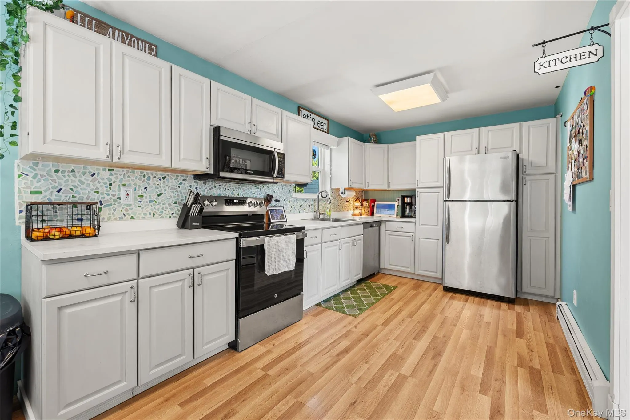Full kitchen featuring white cabinetry, light-toned countertops, a mosaic tile backsplash, stainless steel appliances, and wood-finish flooring Full kitchen featuring white cabinetry, light-toned countertops, a mosaic tile backsplash, stainless steel appliances, and wood-finish flooring