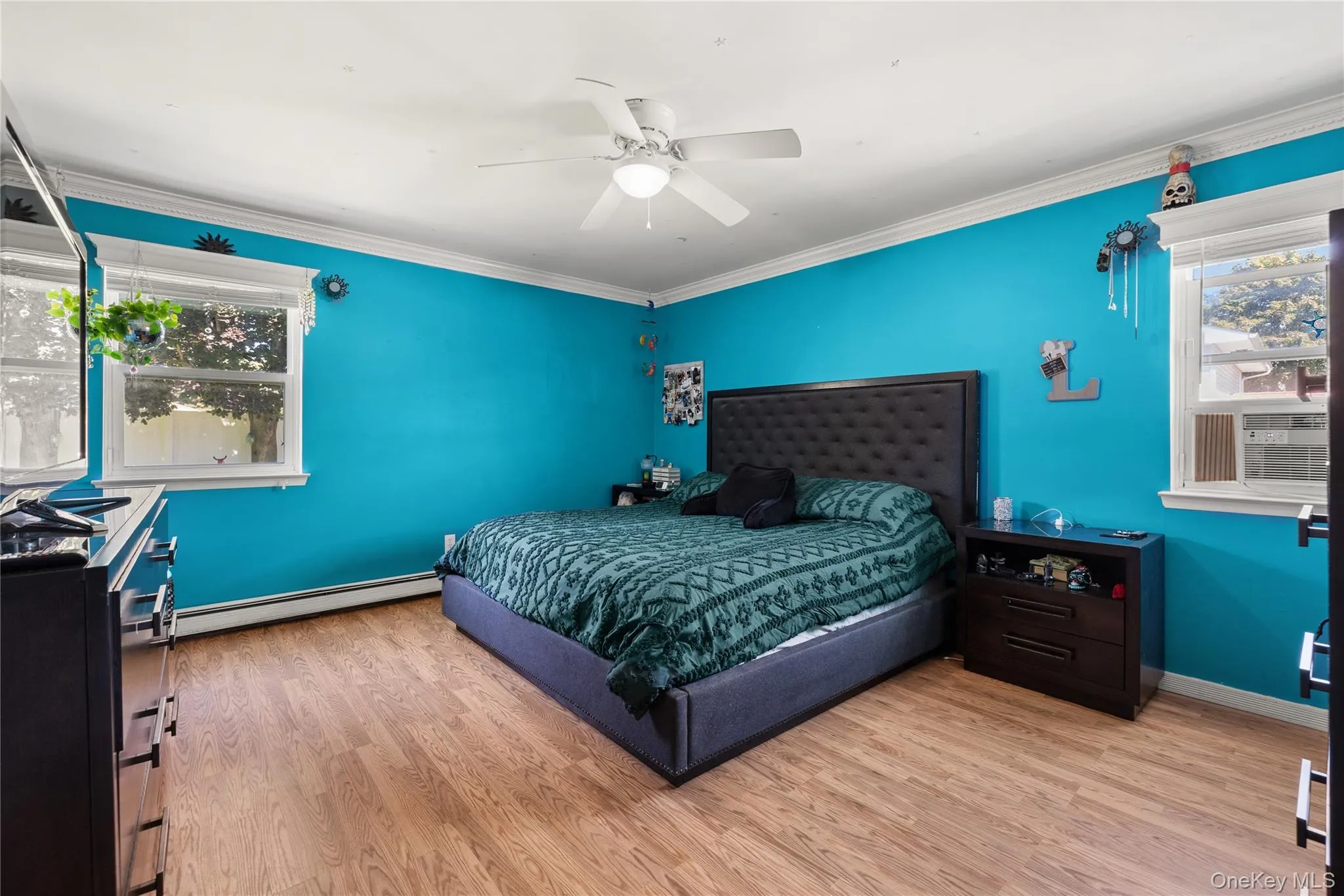 Spacious primary bedroom featuring vibrant turquoise walls, wood-finish flooring, and abundant natural light from dual windows Spacious primary bedroom featuring vibrant turquoise walls, wood-finish flooring, and abundant natural light from dual windows