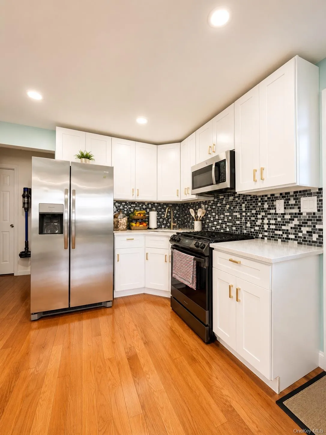 Kitchen featuring white shaker cabinetry with gold-tone hardware, stainless steel appliances, a black and white mosaic tile backsplash, and light wood-finish flooring Kitchen featuring white shaker cabinetry with gold-tone hardware, stainless steel appliances, a black and white mosaic tile backsplash, and light wood-finish flooring