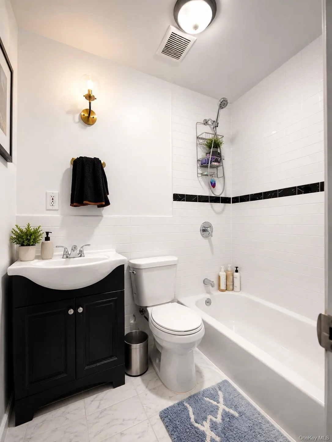 Full bathroom featuring a white tile shower with a dark accent band, a white vanity sink with a black base, a white toilet, and light-toned floor tiling Full bathroom featuring a white tile shower with a dark accent band, a white vanity sink with a black base, a white toilet, and light-toned floor tiling