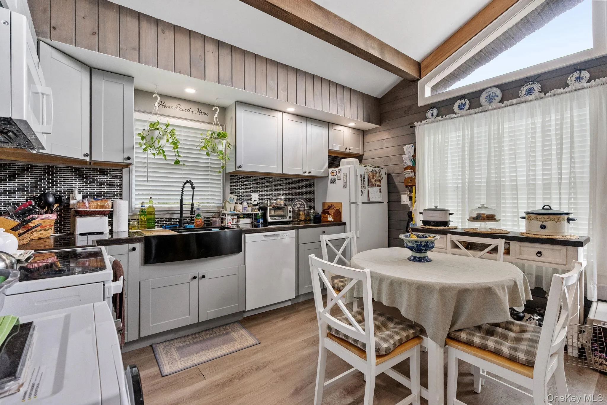 Kitchen with light wood-type flooring, white appliances, wooden walls, dark countertops, and decorative backsplash Kitchen with light wood-type flooring, white appliances, wooden walls, dark countertops, and decorative backsplash