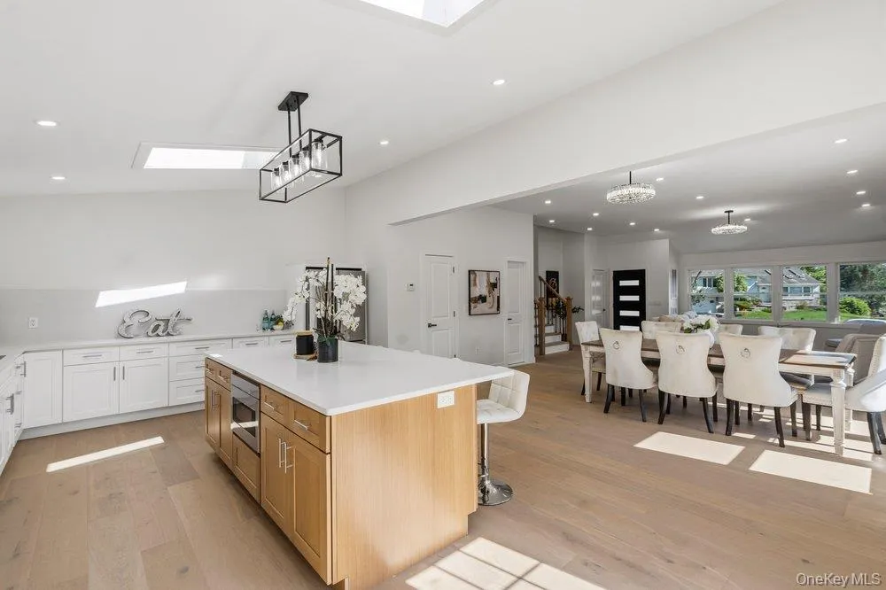 Expansive kitchen and dining area featuring light wood-finish flooring, a kitchen island with built-in appliances and a light-toned countertop, white cabinetry with silver hardware, and recessed ceiling lighting Expansive kitchen and dining area featuring light wood-finish flooring, a kitchen island with built-in appliances and a light-toned countertop, white cabinetry with silver hardware, and recessed ceiling lighting