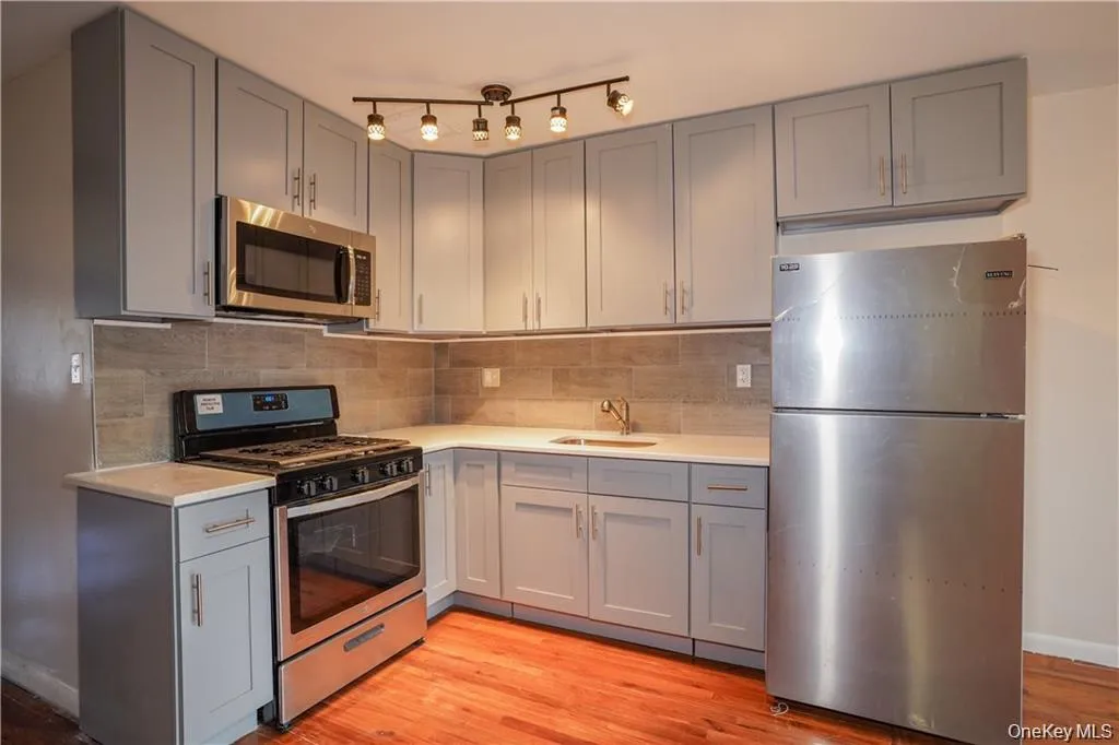 Kitchen featuring light gray cabinetry, stainless steel appliances, a tile backsplash, light-colored countertops, and hardwood flooring Kitchen featuring light gray cabinetry, stainless steel appliances, a tile backsplash, light-colored countertops, and hardwood flooring