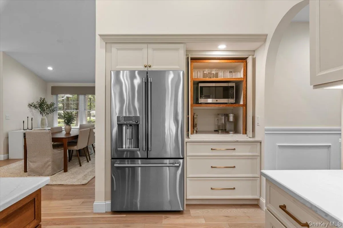 Stainless steel French door refrigerator, integrated microwave cabinet with wood-finish interior, light-toned wood-finish flooring, white kitchen cabinetry with bronze-finish hardware, and a white countertop Stainless steel French door refrigerator, integrated microwave cabinet with wood-finish interior, light-toned wood-finish flooring, white kitchen cabinetry with bronze-finish hardware, and a white countertop