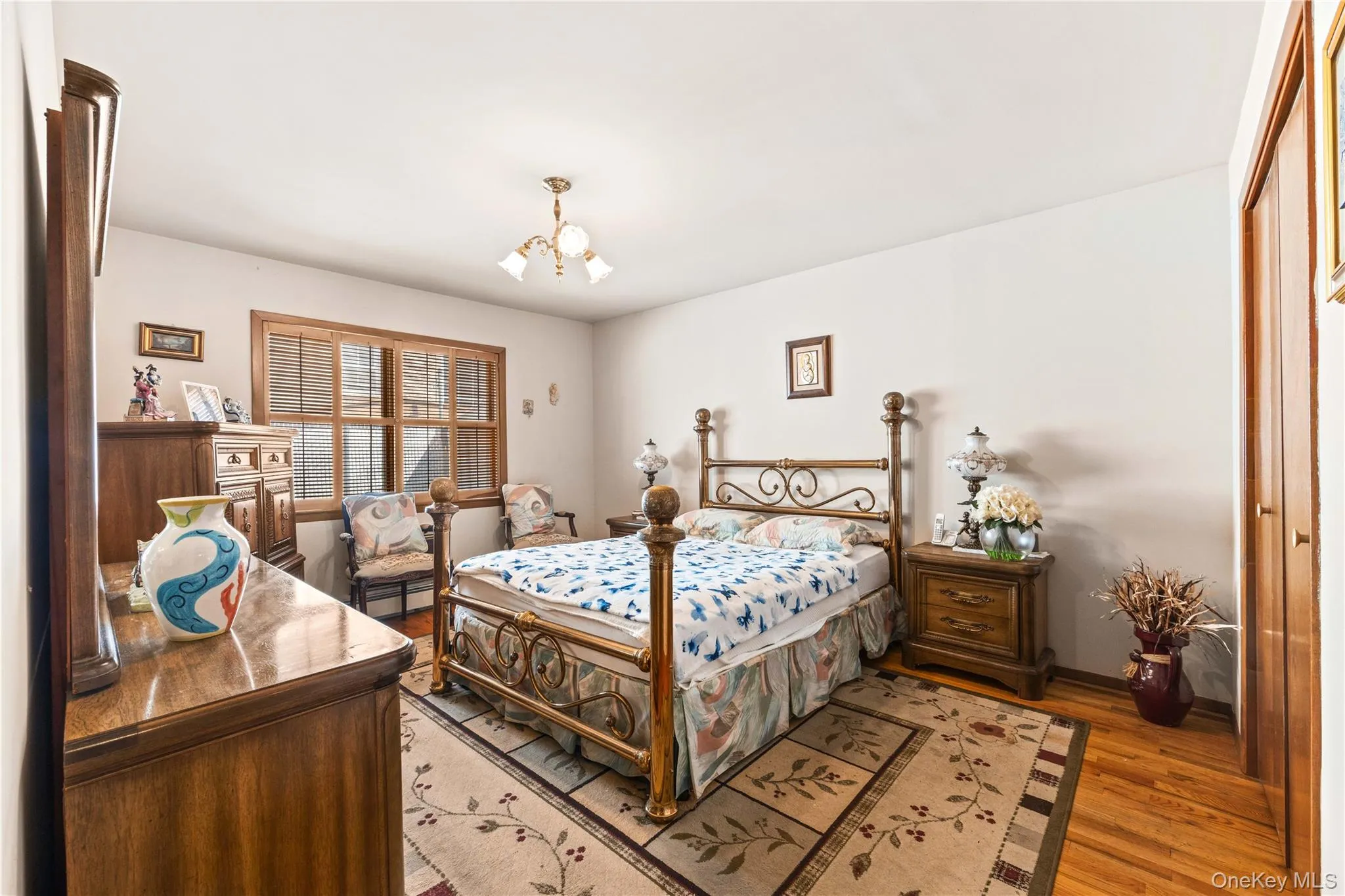 Bedroom featuring wood-finish flooring, a multi-pane window with interior shutters, and a brass-tone chandelier Bedroom featuring wood-finish flooring, a multi-pane window with interior shutters, and a brass-tone chandelier