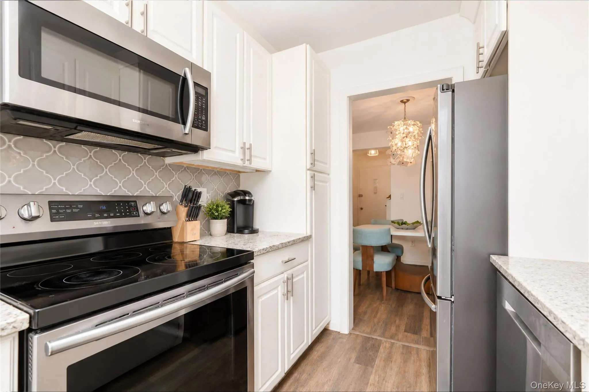 Kitchen featuring white shaker-style cabinetry with brushed nickel hardware, stainless steel appliances, light-toned countertops, and a gray arabesque tile backsplash Kitchen featuring white shaker-style cabinetry with brushed nickel hardware, stainless steel appliances, light-toned countertops, and a gray arabesque tile backsplash