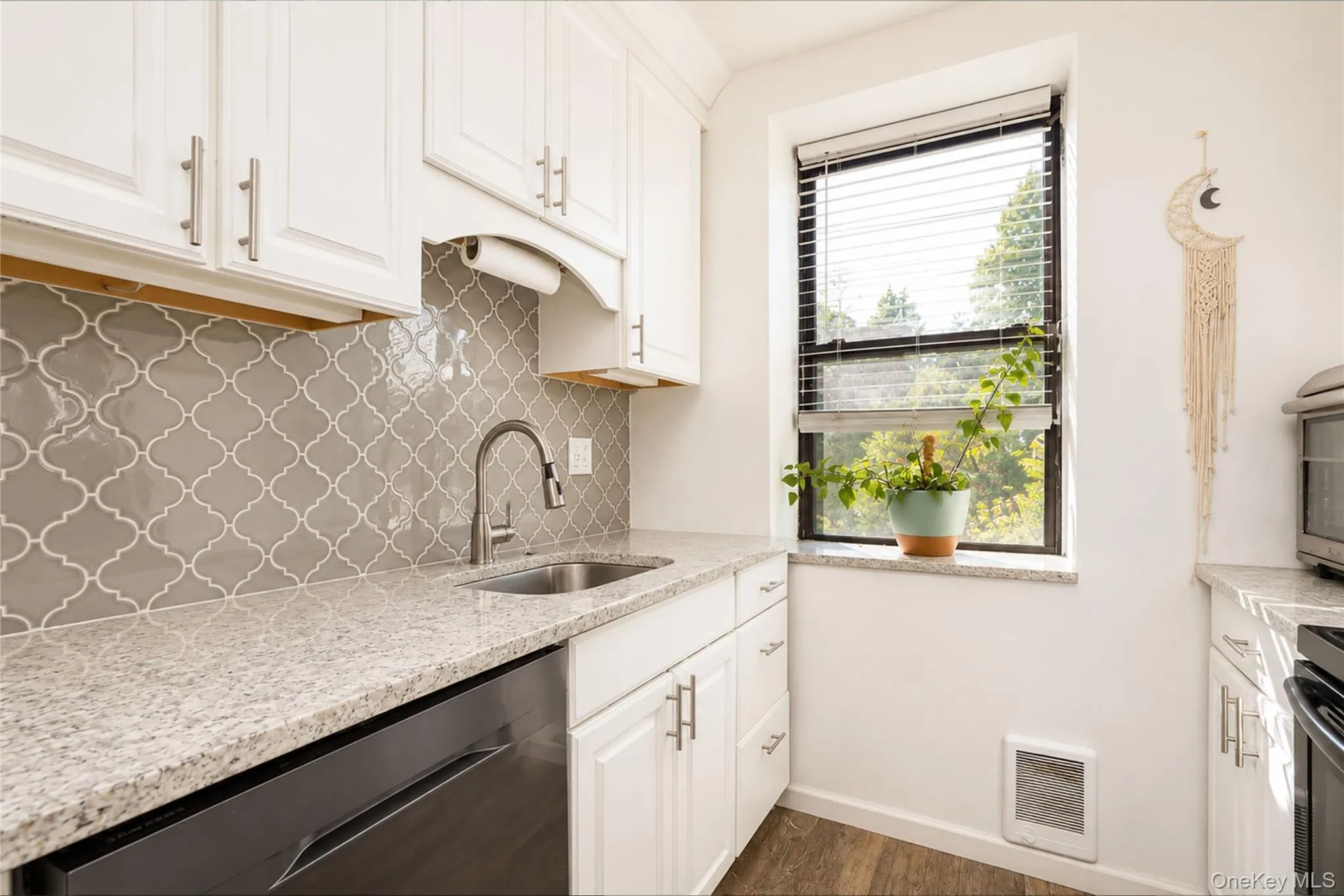 Kitchen featuring white upper and lower cabinetry with bar pull hardware, a stainless steel sink with a gooseneck faucet, light-toned granite countertops, and a gray arabesque tile backsplash Kitchen featuring white upper and lower cabinetry with bar pull hardware, a stainless steel sink with a gooseneck faucet, light-toned granite countertops, and a gray arabesque tile backsplash