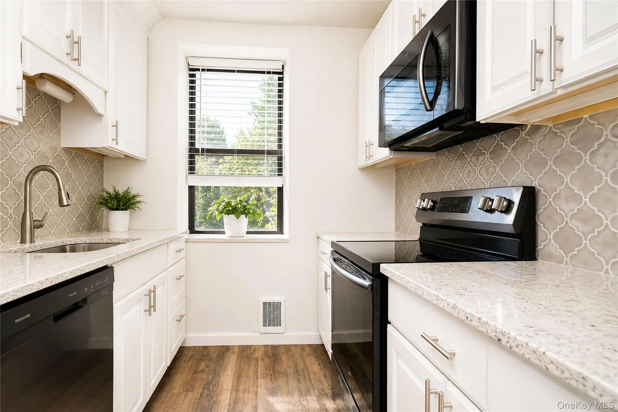 Galley kitchen featuring white cabinetry with brushed nickel hardware, quartz countertops, geometric tile backsplash, and stainless steel appliances including a black range and microwave Galley kitchen featuring white cabinetry with brushed nickel hardware, quartz countertops, geometric tile backsplash, and stainless steel appliances including a black range and microwave