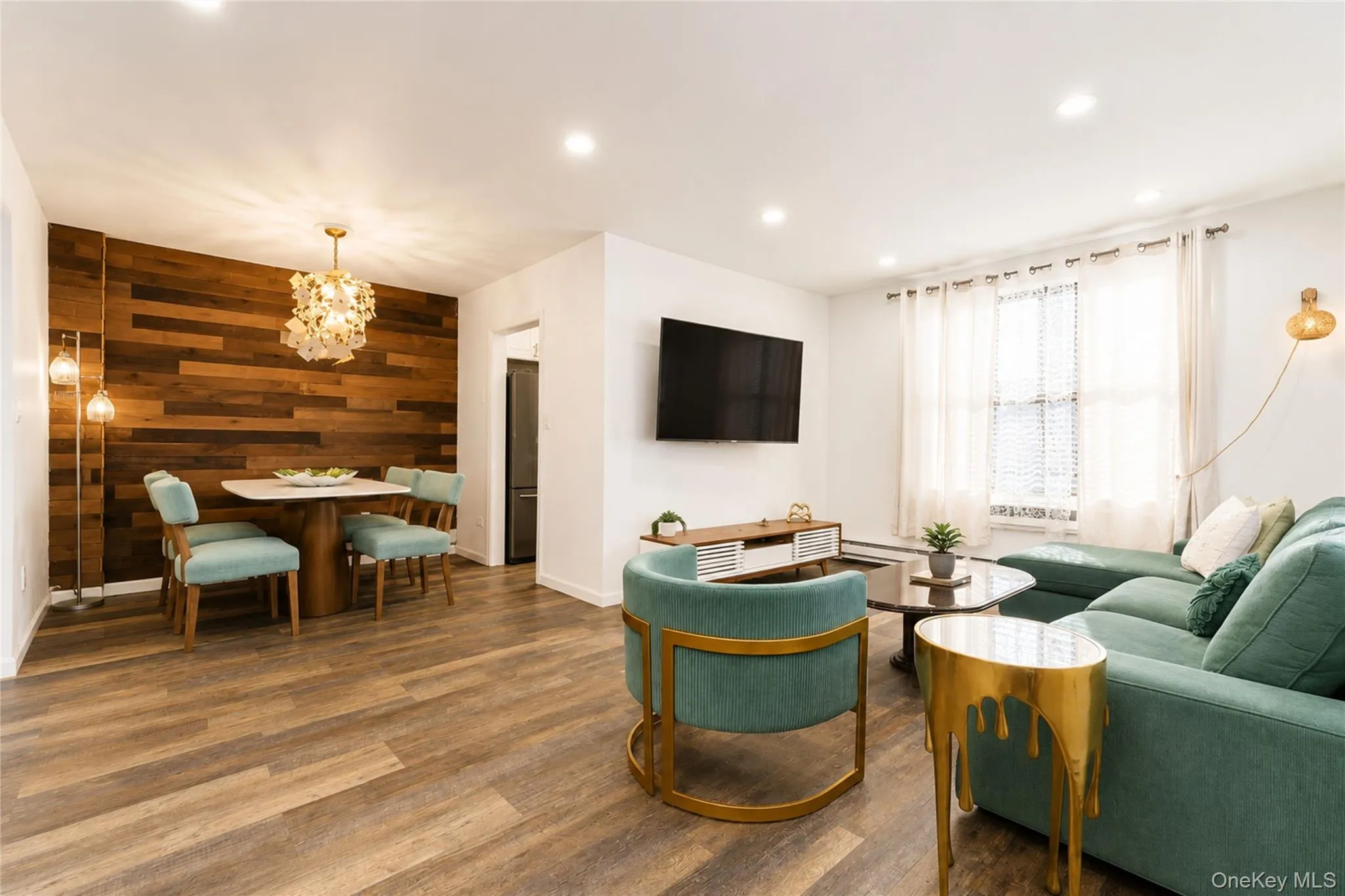 Open-concept living area featuring wood-finish flooring, a dining space with a unique decorative chandelier, and a prominent wood-panel accent wall Open-concept living area featuring wood-finish flooring, a dining space with a unique decorative chandelier, and a prominent wood-panel accent wall