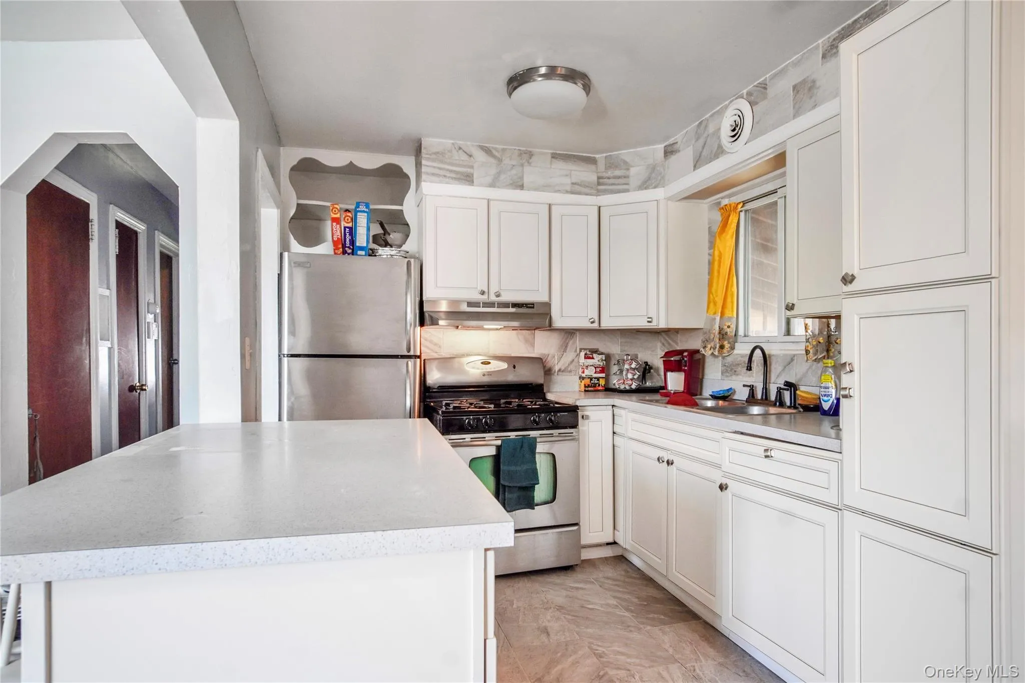 Bright kitchen featuring white cabinetry, a white countertop island, stainless steel appliances, and a marble-patterned tile backsplash Bright kitchen featuring white cabinetry, a white countertop island, stainless steel appliances, and a marble-patterned tile backsplash