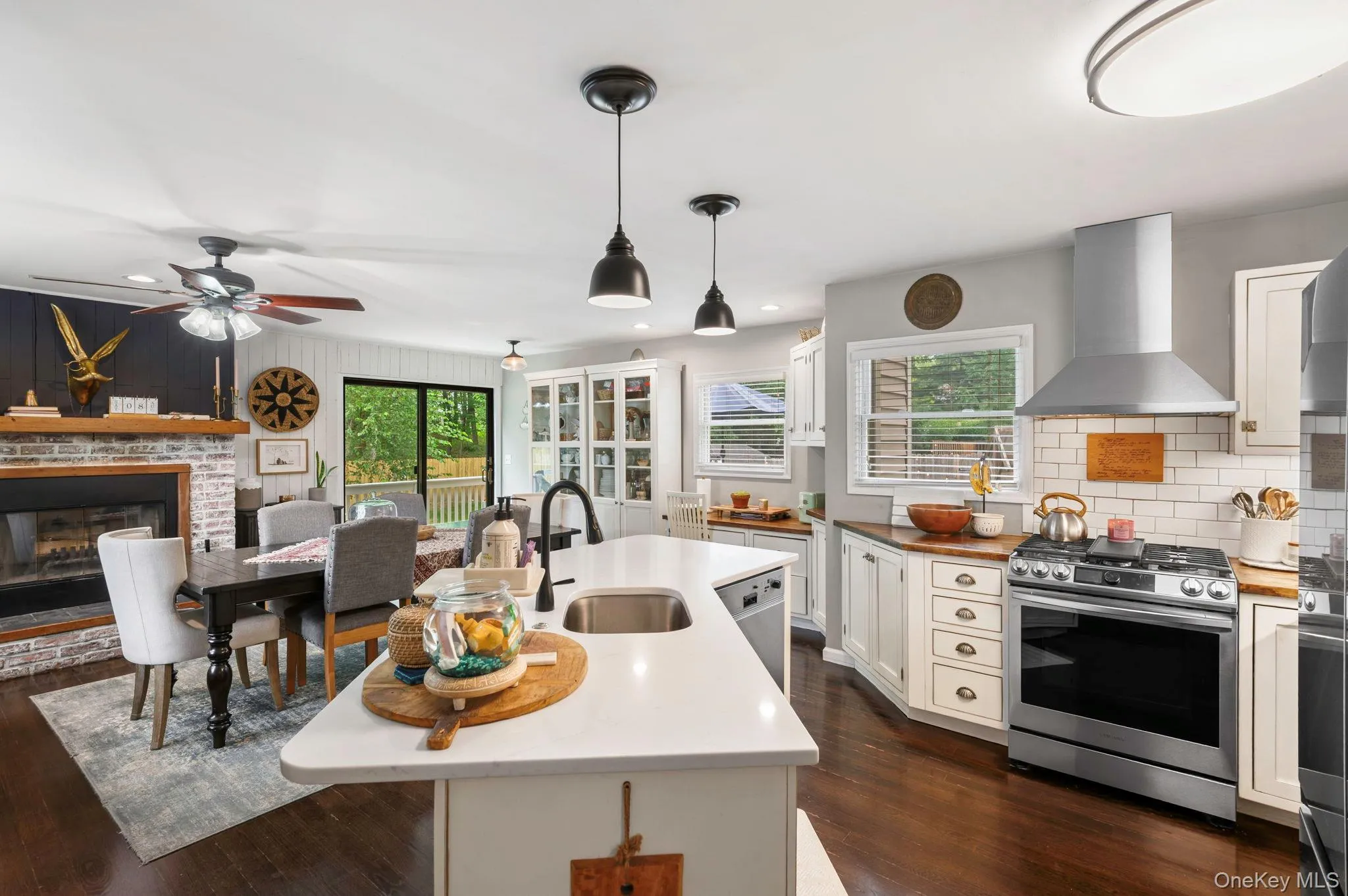Open-concept kitchen featuring an island with a sink, wood-finish flooring, white cabinetry, a stainless steel range with a hood, and a brick fireplace Open-concept kitchen featuring an island with a sink, wood-finish flooring, white cabinetry, a stainless steel range with a hood, and a brick fireplace