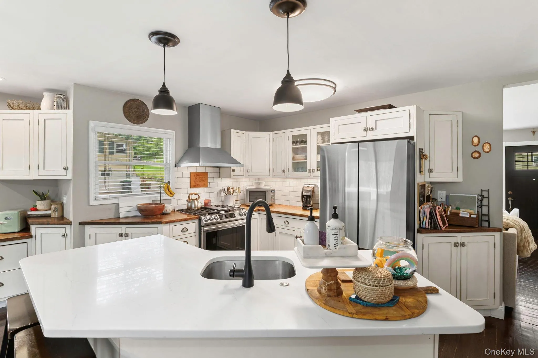 Kitchen featuring white cabinetry, a stainless steel range hood, a stainless steel refrigerator, white subway tile backsplash, and a large central island with an undermount sink Kitchen featuring white cabinetry, a stainless steel range hood, a stainless steel refrigerator, white subway tile backsplash, and a large central island with an undermount sink