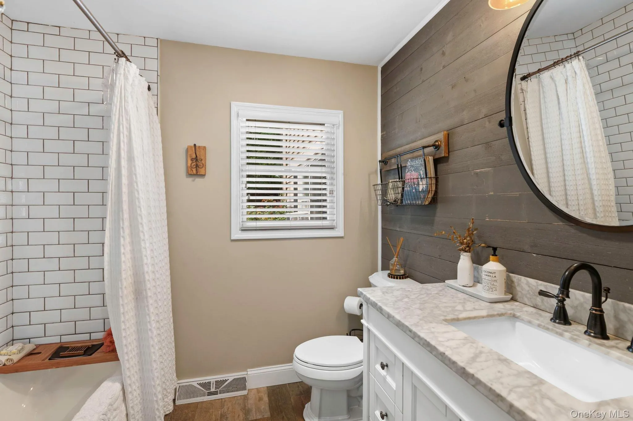 Subway tile shower surround, window with blinds, wood-finish accent wall, white vanity with stone countertop, and dark-finish fixtures Subway tile shower surround, window with blinds, wood-finish accent wall, white vanity with stone countertop, and dark-finish fixtures