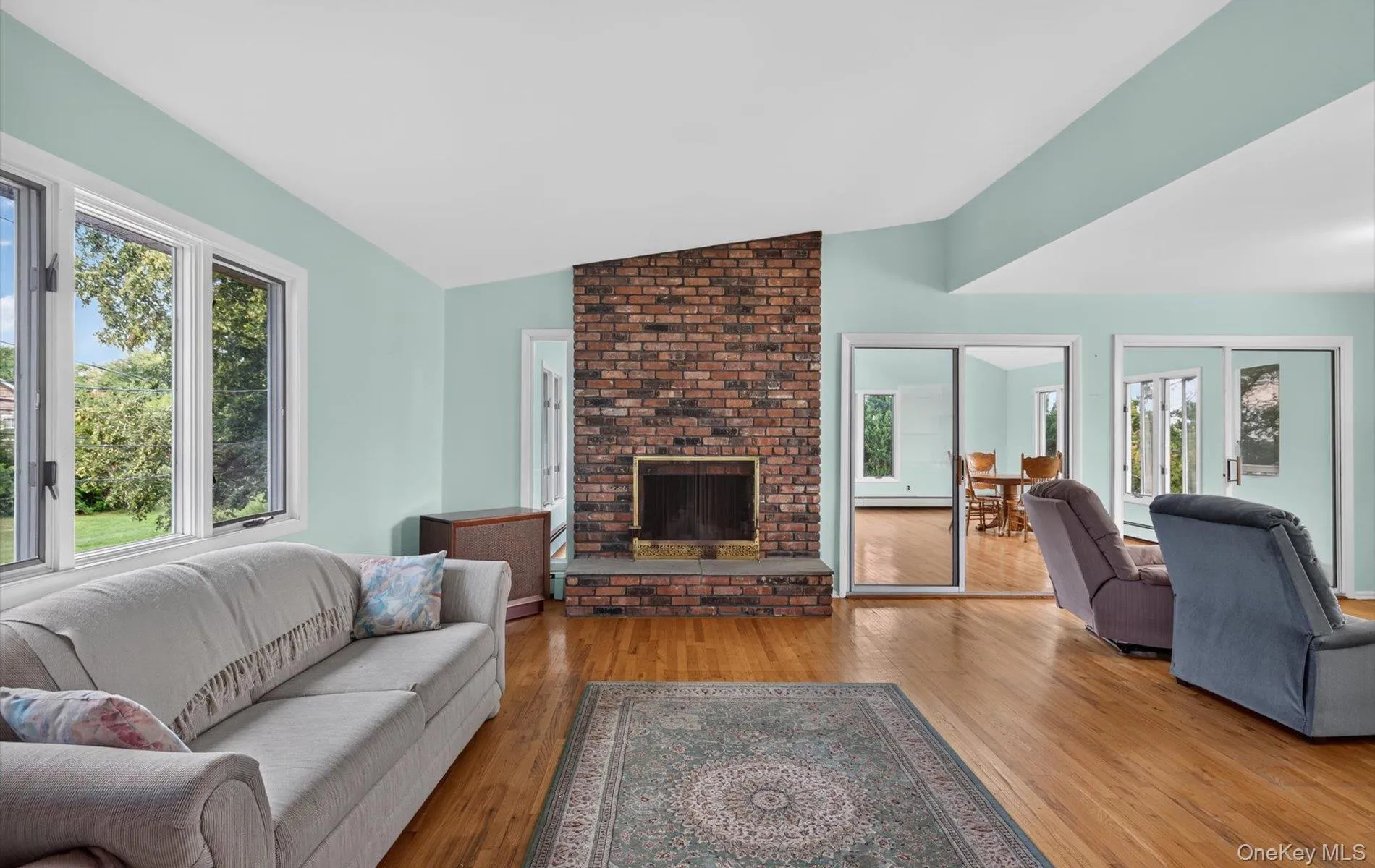 Brick fireplace with hearth, wood-finish flooring, multiple windows, and sliding glass doors Brick fireplace with hearth, wood-finish flooring, multiple windows, and sliding glass doors