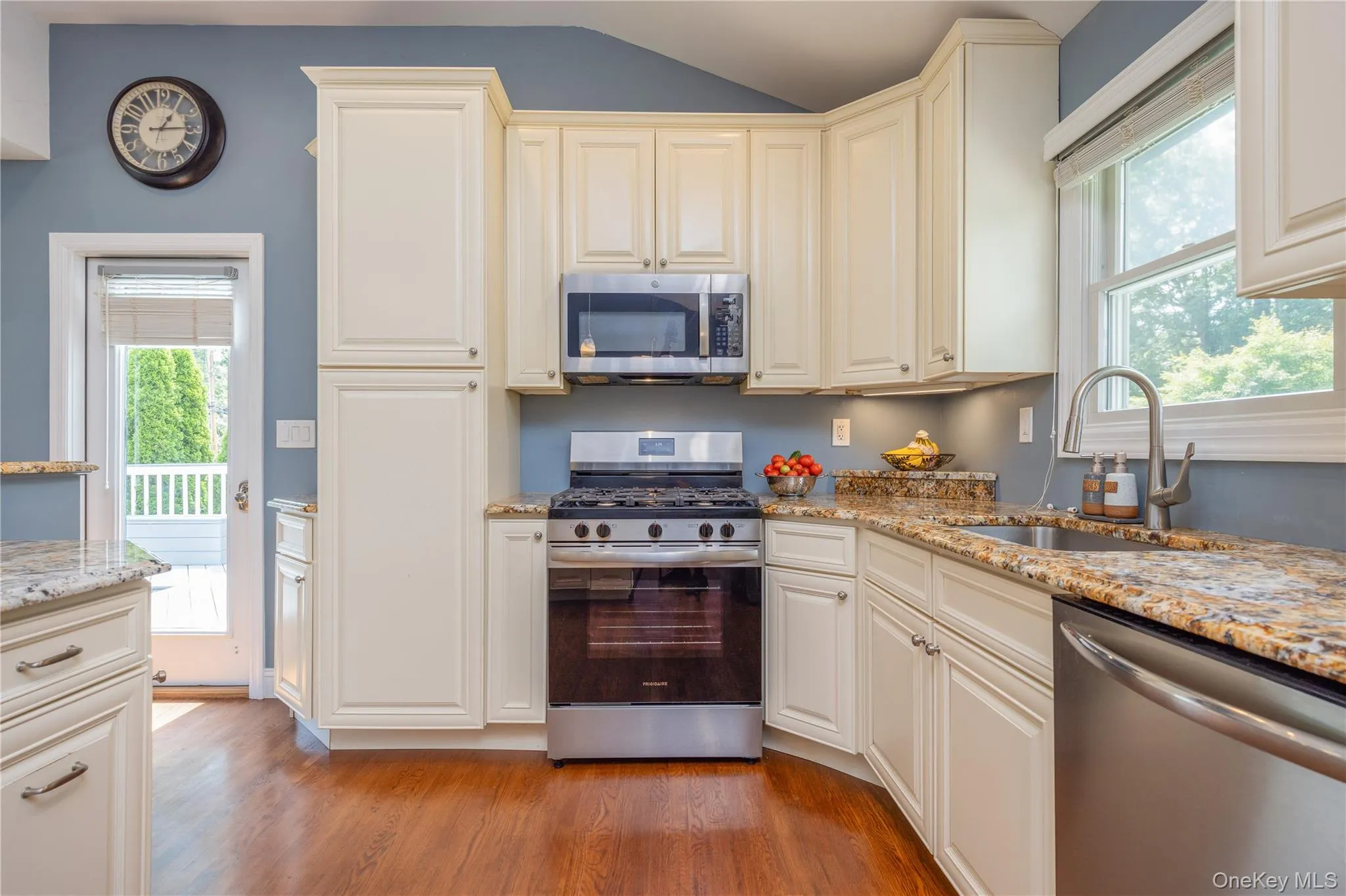 Elegant kitchen featuring cream-colored cabinetry with crown molding, granite countertops, stainless steel appliances including a gas range and built-in microwave, hardwood flooring, and abundant natural light from multiple windows Elegant kitchen featuring cream-colored cabinetry with crown molding, granite countertops, stainless steel appliances including a gas range and built-in microwave, hardwood flooring, and abundant natural light from multiple windows