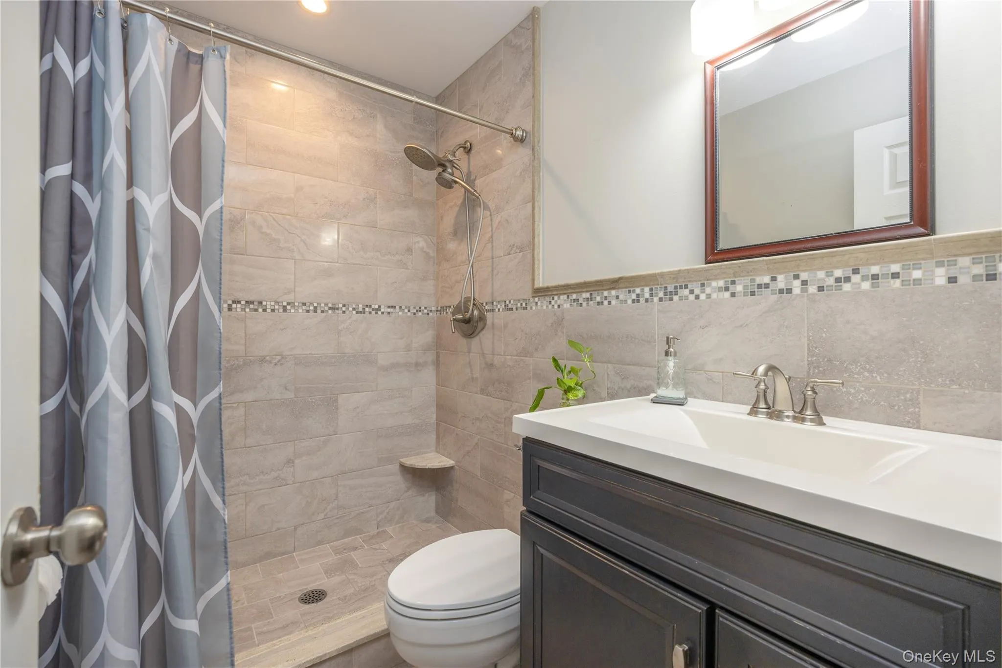 Modern bathroom featuring a spacious walk-in shower with tile surround and glass enclosure, a sleek vanity with white quartz countertop and dual-sink configuration, brushed nickel fixtures, and mosaic tile accent band Modern bathroom featuring a spacious walk-in shower with tile surround and glass enclosure, a sleek vanity with white quartz countertop and dual-sink configuration, brushed nickel fixtures, and mosaic tile accent band