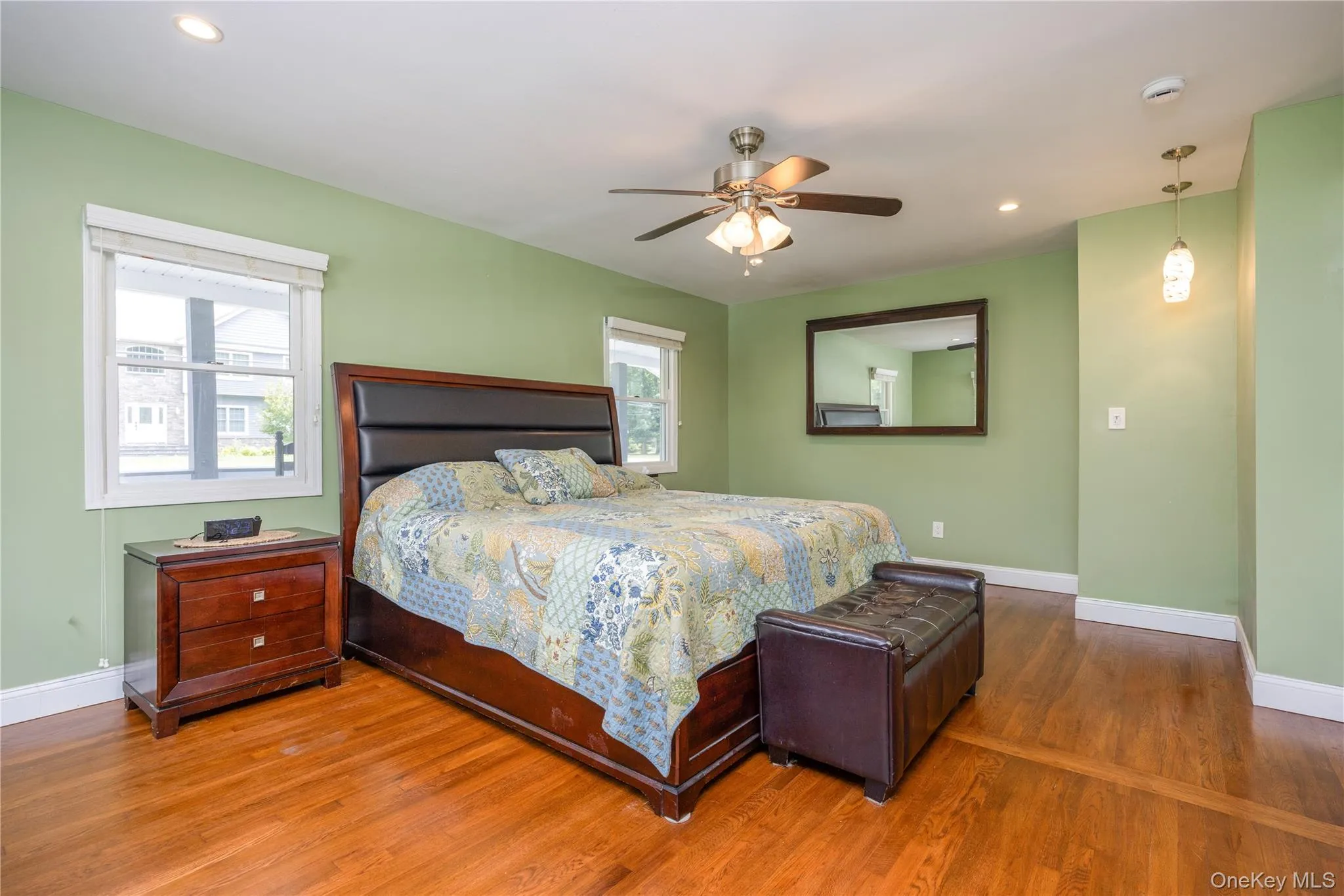 Spacious primary bedroom showcasing soft green walls, hardwood flooring, ceiling fan with integrated lighting, and a framed mirror Spacious primary bedroom showcasing soft green walls, hardwood flooring, ceiling fan with integrated lighting, and a framed mirror