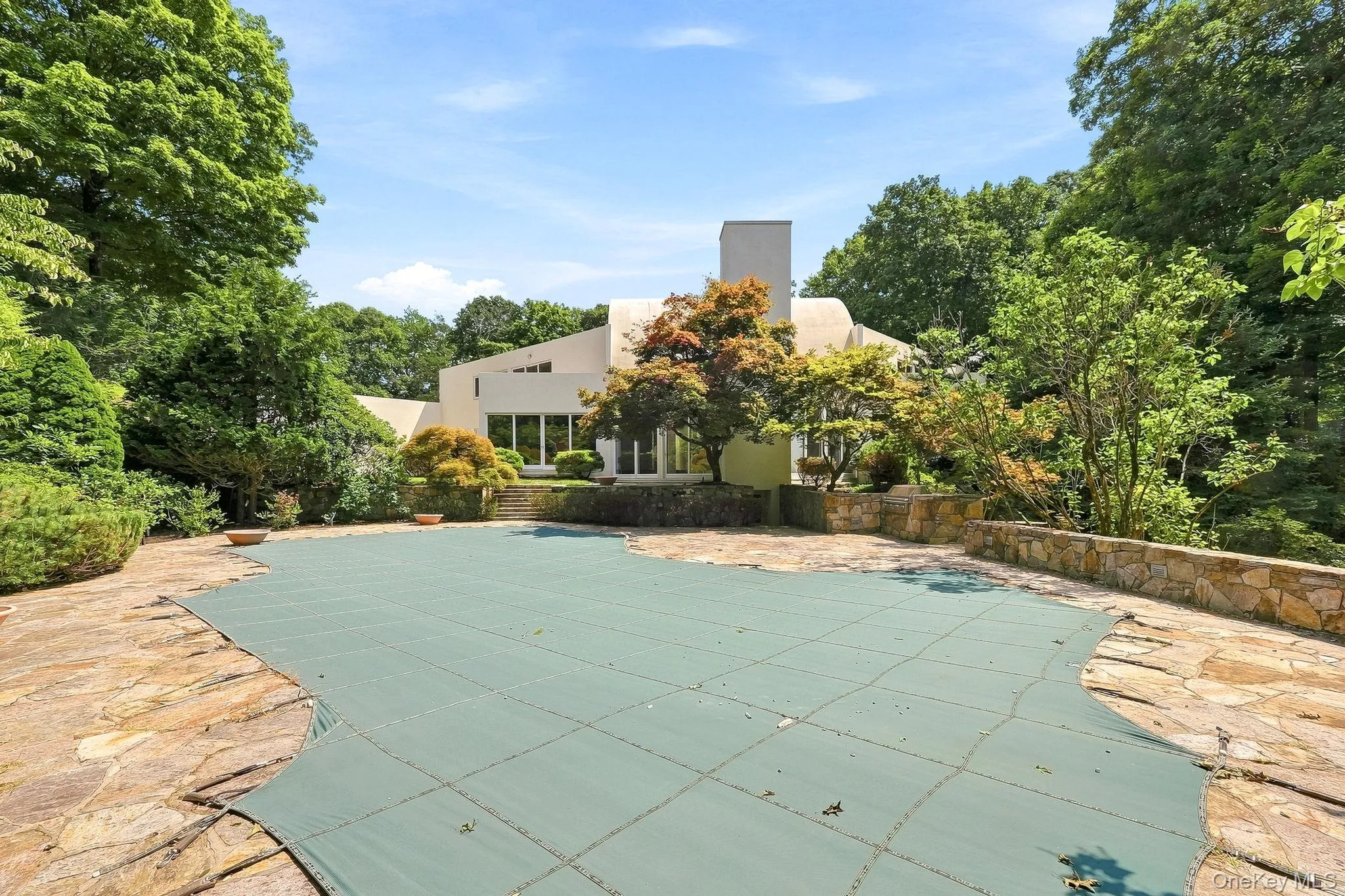 An expansive stone patio surrounds a private, tree-lined swimming pool overlooked by the striking modern estate facade. An expansive stone patio surrounds a private, tree-lined swimming pool overlooked by the striking modern estate facade.