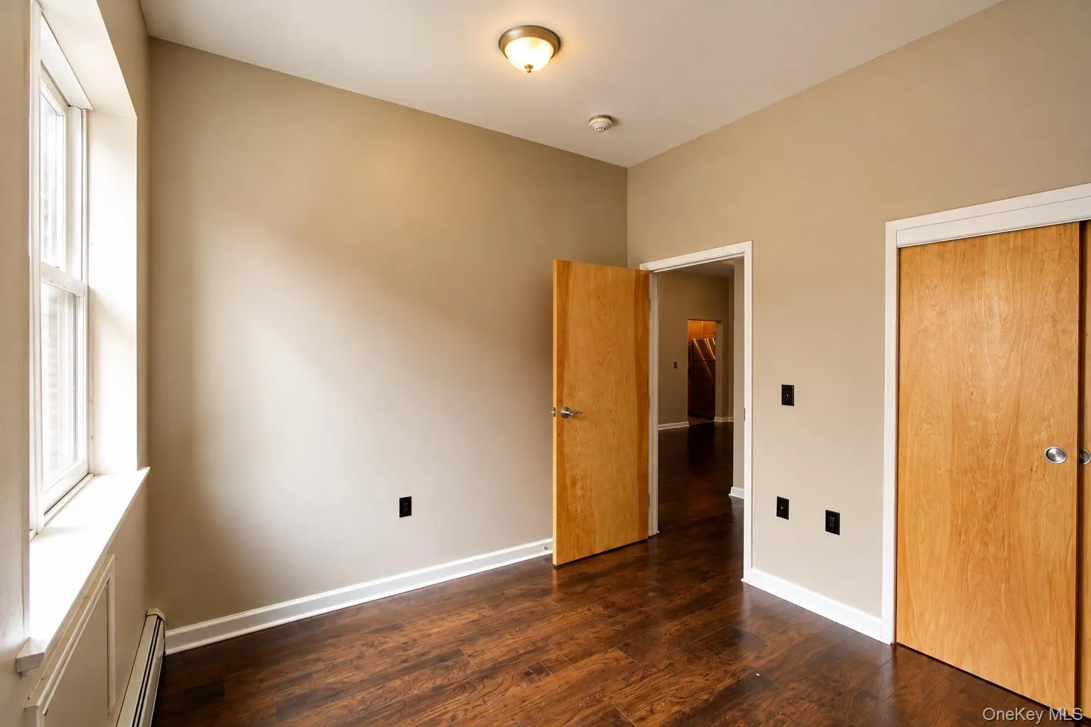 Spacious bedroom with rich wood-finish flooring, abundant natural light from large window, and multiple closet entries with wood-finish doors Spacious bedroom with rich wood-finish flooring, abundant natural light from large window, and multiple closet entries with wood-finish doors
