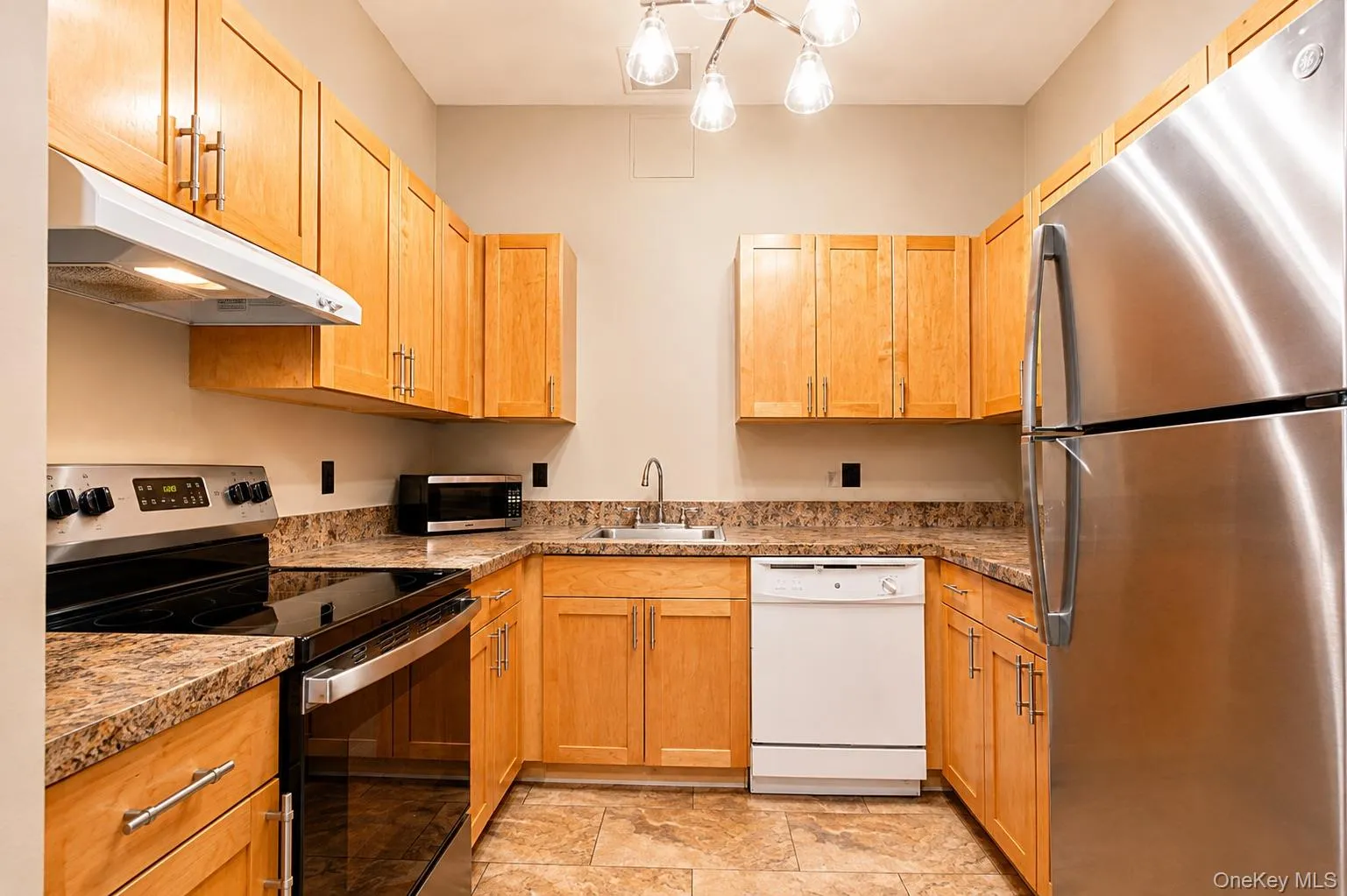 U-shaped kitchen with wood cabinetry, speckled countertops, stainless steel refrigerator, white dishwasher, and dark tile flooring U-shaped kitchen with wood cabinetry, speckled countertops, stainless steel refrigerator, white dishwasher, and dark tile flooring