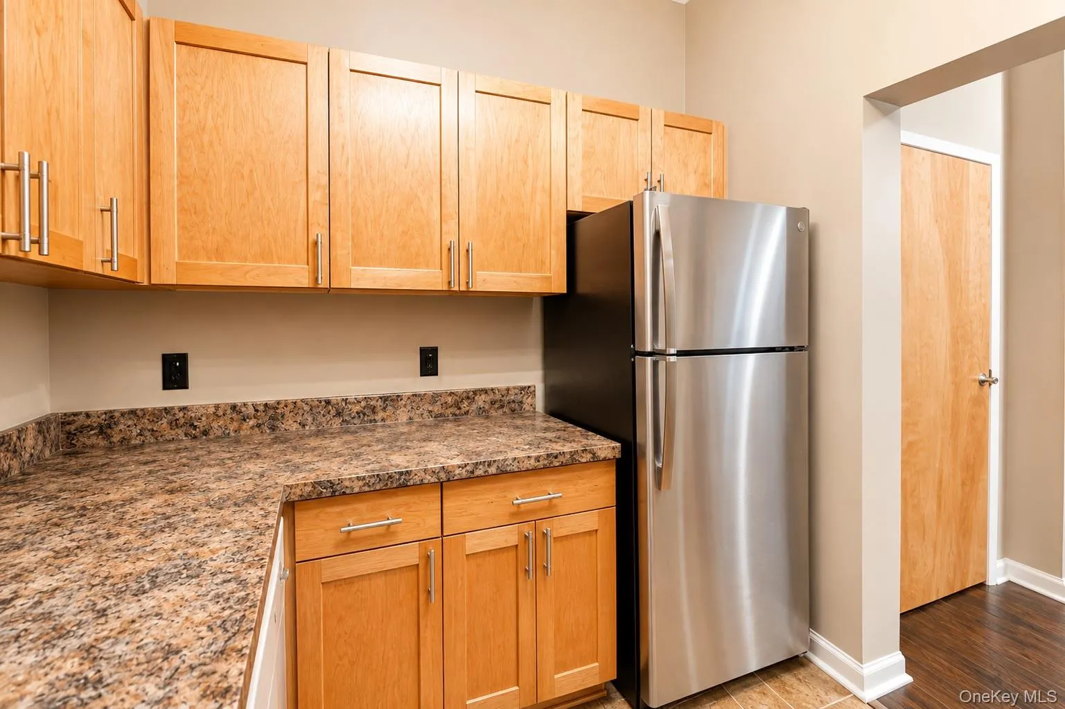 Wood-finish cabinetry, speckled countertops, stainless steel refrigerator, and wood-finish flooring Wood-finish cabinetry, speckled countertops, stainless steel refrigerator, and wood-finish flooring