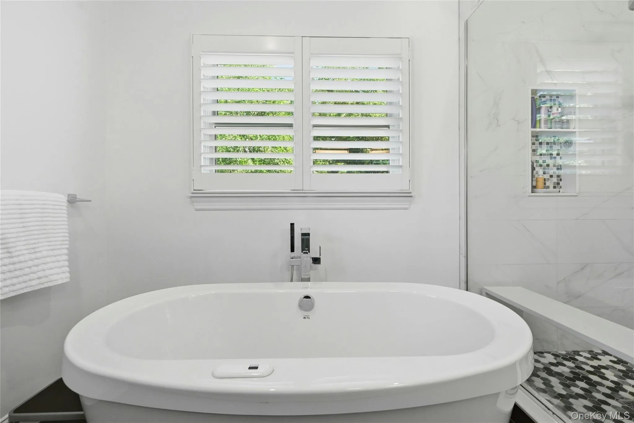 Spa-inspired primary bath featuring a freestanding oval soaking tub, marble-look wall tile surround, polished stone-look flooring with geometric mosaic accent, and plantation shutters Spa-inspired primary bath featuring a freestanding oval soaking tub, marble-look wall tile surround, polished stone-look flooring with geometric mosaic accent, and plantation shutters