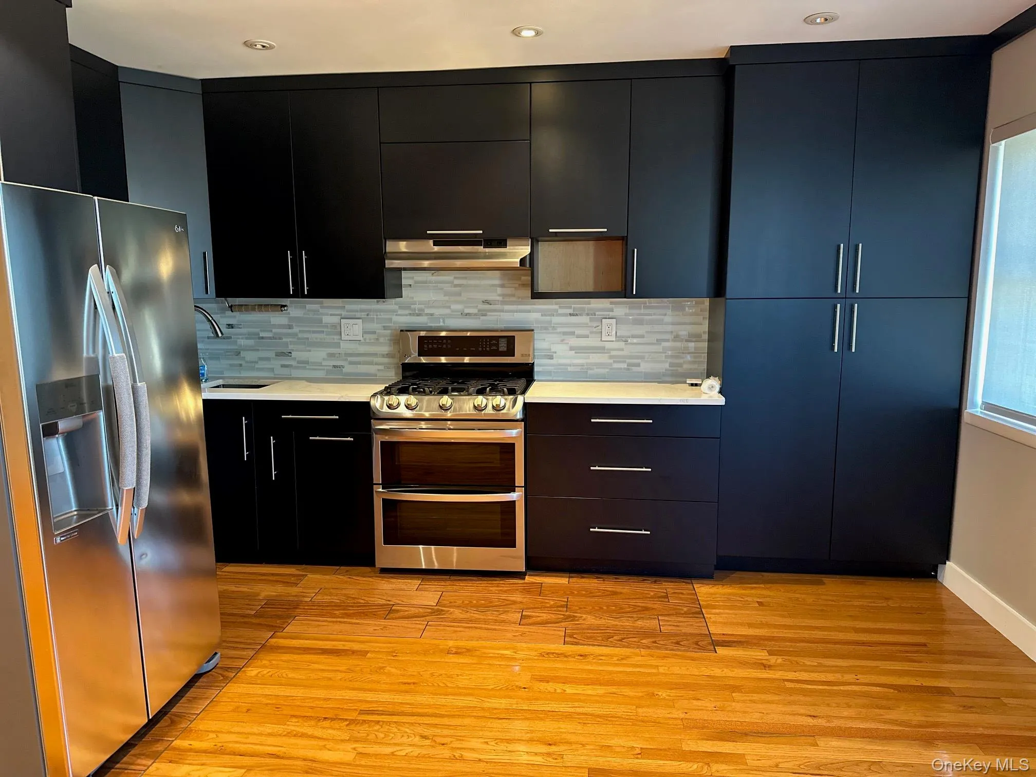 Kitchen featuring black flat-panel cabinetry, a tile backsplash, stainless steel appliances, light-colored countertops, and wood flooring Kitchen featuring black flat-panel cabinetry, a tile backsplash, stainless steel appliances, light-colored countertops, and wood flooring