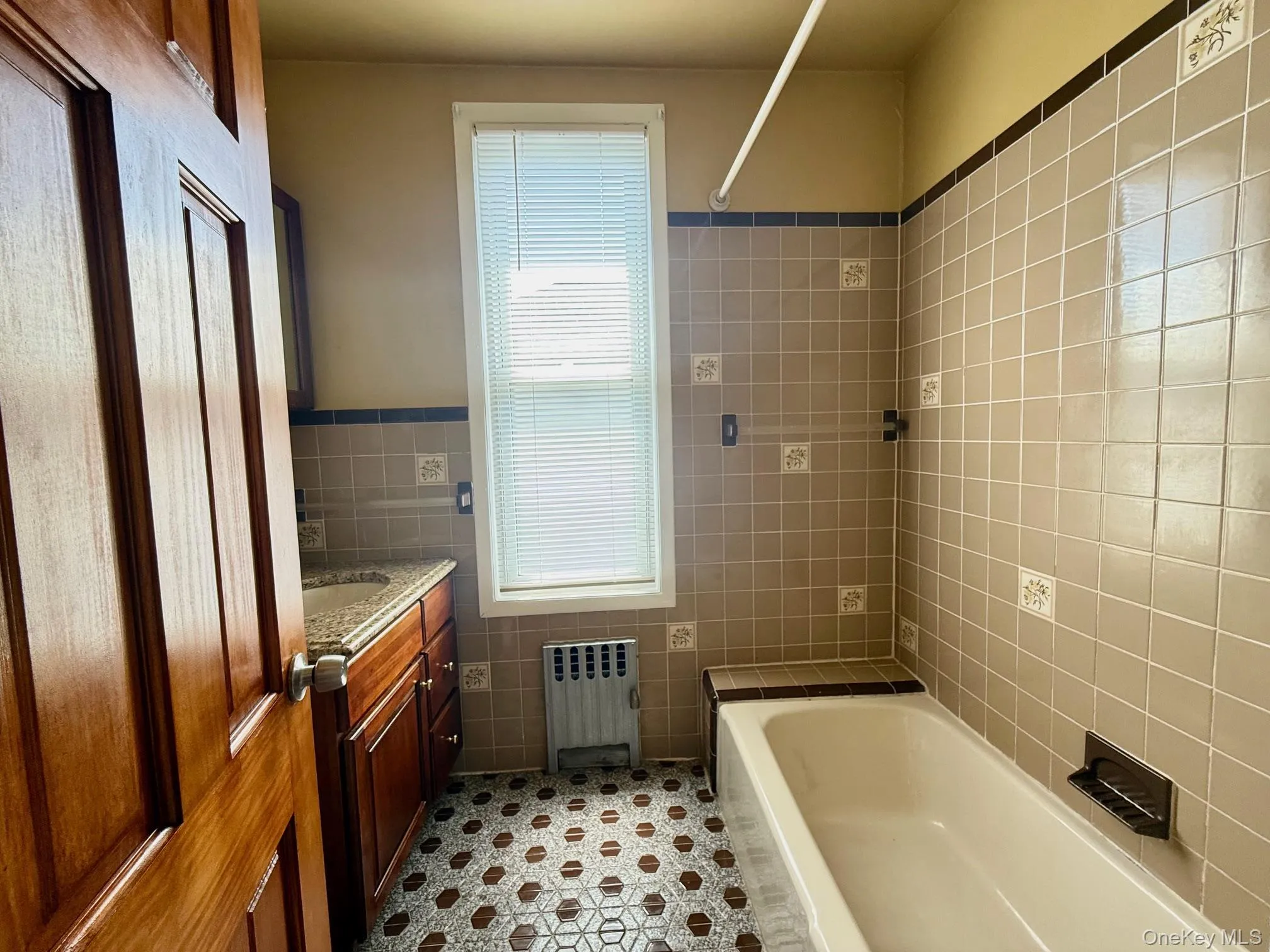 Tiled bathroom featuring a built-in bathtub, a window with blinds, and a vanity with a countertop and under-sink cabinetry Tiled bathroom featuring a built-in bathtub, a window with blinds, and a vanity with a countertop and under-sink cabinetry
