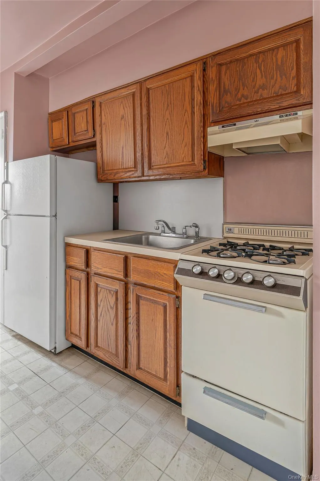 Kitchenette featuring wood-finish cabinetry, a built-in sink, a freestanding refrigerator, a gas range, and square tile flooring Kitchenette featuring wood-finish cabinetry, a built-in sink, a freestanding refrigerator, a gas range, and square tile flooring