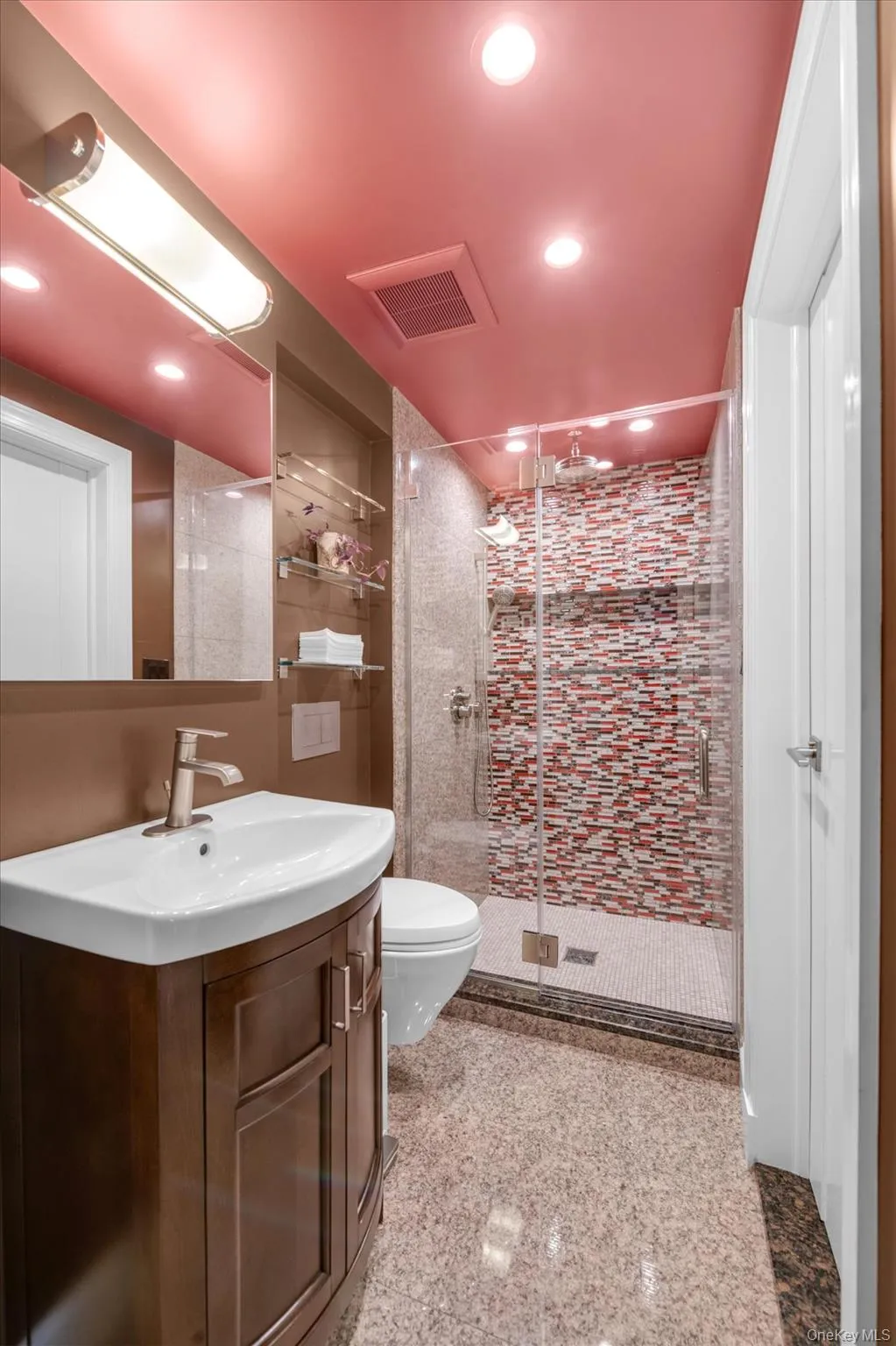 Spa-inspired bathroom featuring ceiling with recessed lighting, vanity with wood cabinetry and white vessel sink, mosaic tile shower enclosure with glass partition, and polished tile flooring throughout Spa-inspired bathroom featuring ceiling with recessed lighting, vanity with wood cabinetry and white vessel sink, mosaic tile shower enclosure with glass partition, and polished tile flooring throughout