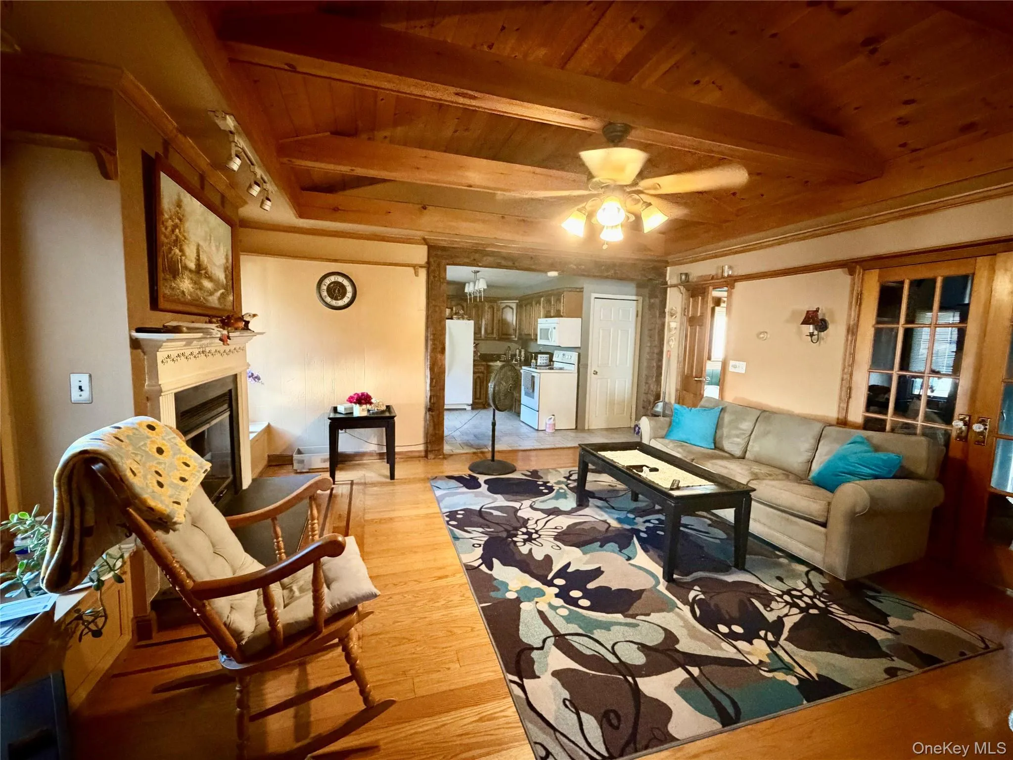Wood-finish flooring, wood plank ceiling with exposed beams, fireplace with decorative mantel, and a ceiling fan with integrated lighting Wood-finish flooring, wood plank ceiling with exposed beams, fireplace with decorative mantel, and a ceiling fan with integrated lighting