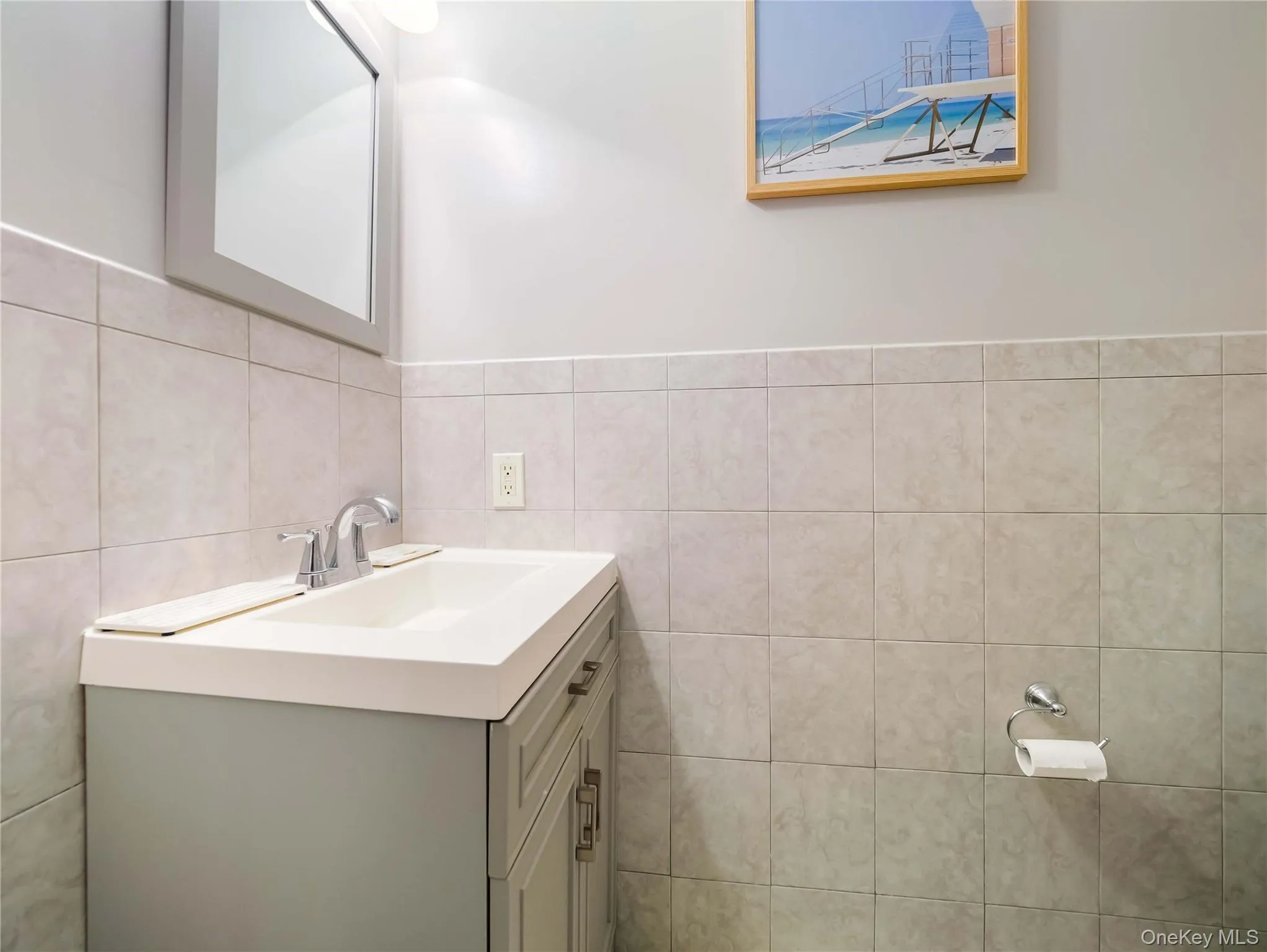 Wall-mounted mirror with gray framing, vanity cabinet with integrated rectangular sink, chrome dual-handle faucet, and tiled wainscoting Wall-mounted mirror with gray framing, vanity cabinet with integrated rectangular sink, chrome dual-handle faucet, and tiled wainscoting