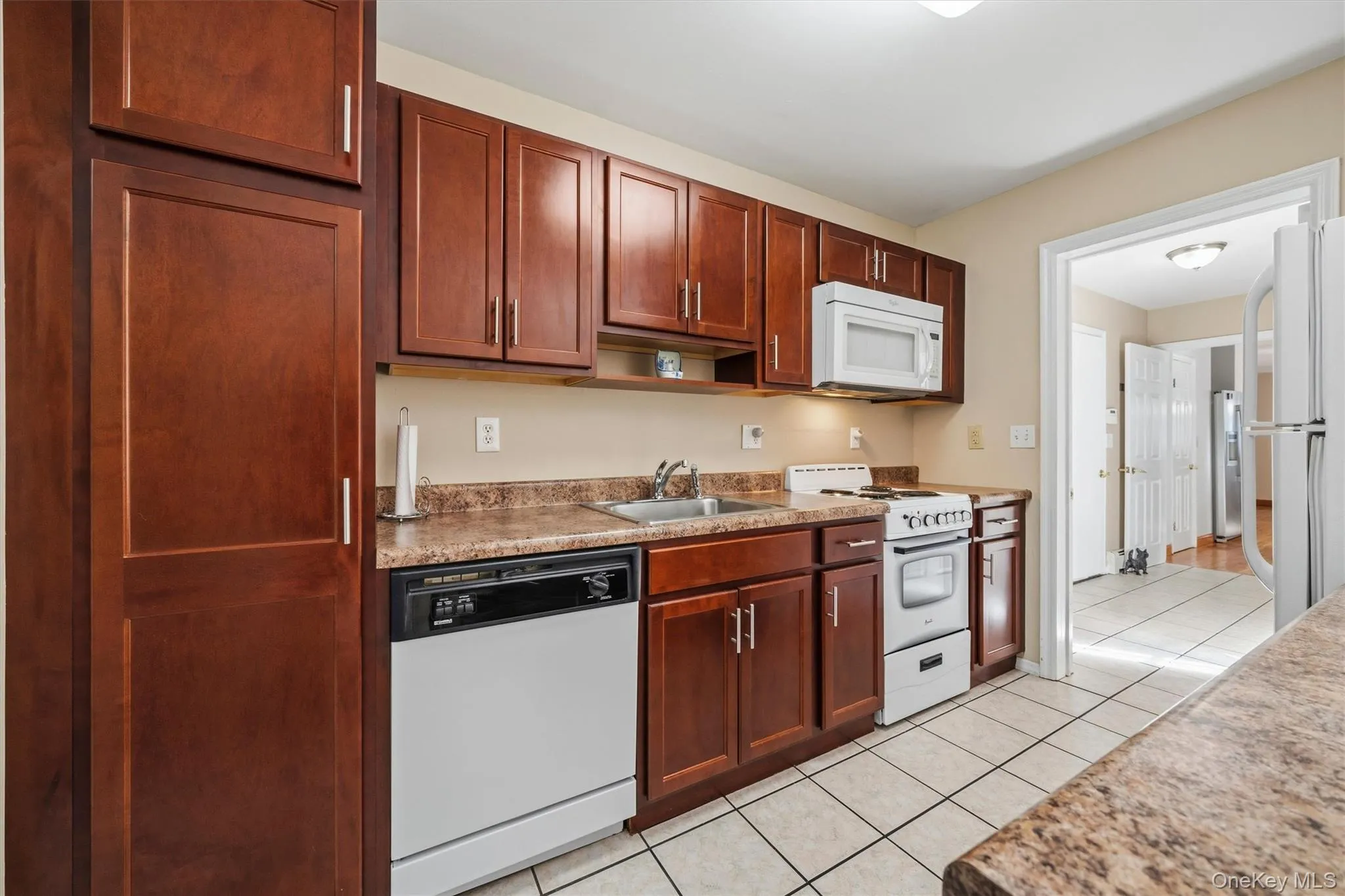 Mother/daughter Kitchen featuring wood-finish cabinetry, speckled countertops, white appliances, and tiled flooring Mother/daughter Kitchen featuring wood-finish cabinetry, speckled countertops, white appliances, and tiled flooring