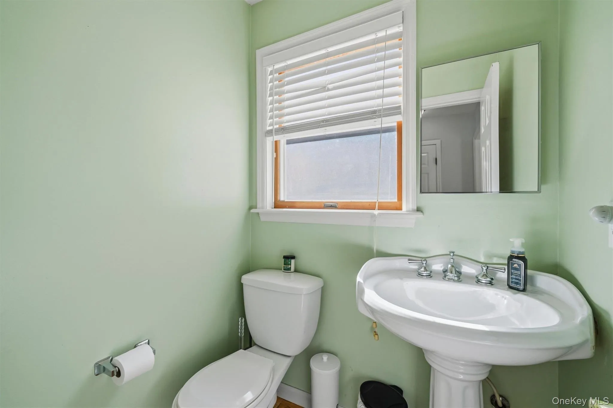Pedestal sink with chrome-finish fixtures, a rectangular window with blinds, and a wall-mounted mirror Pedestal sink with chrome-finish fixtures, a rectangular window with blinds, and a wall-mounted mirror