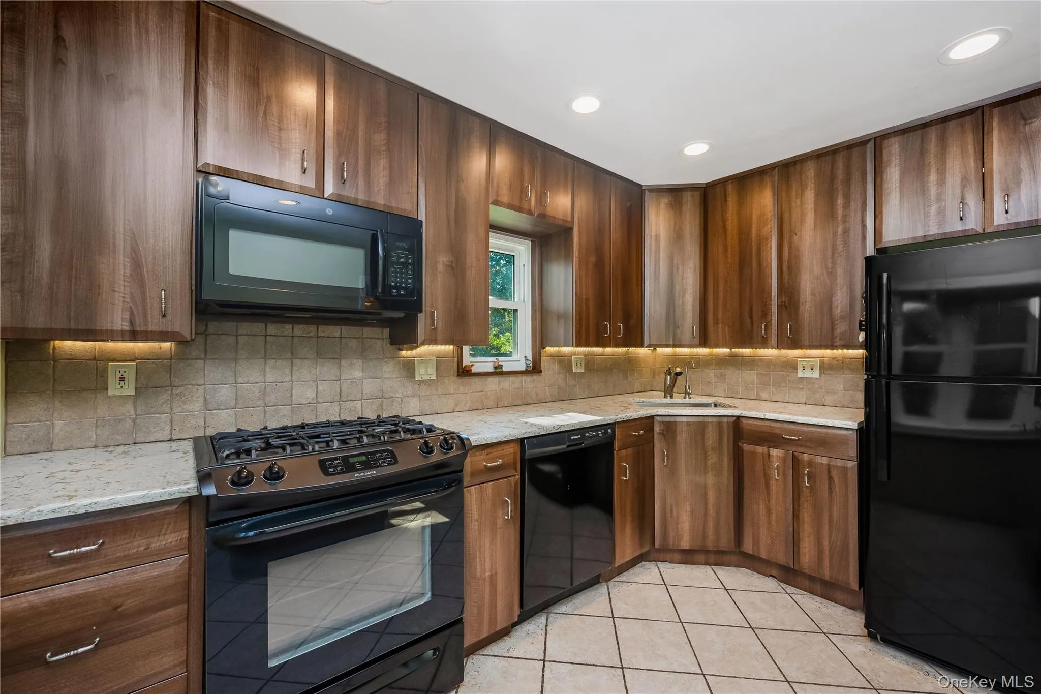 Kitchen featuring wood-finish cabinetry, light-toned countertop, tile backsplash, and black appliances Kitchen featuring wood-finish cabinetry, light-toned countertop, tile backsplash, and black appliances