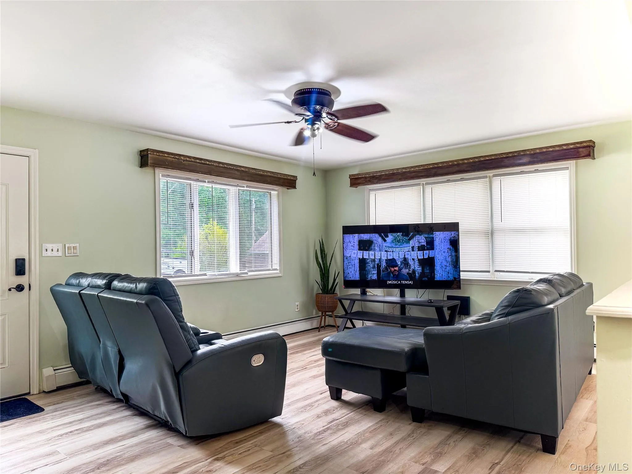 Spacious living room featuring wood-finish flooring, pale green walls, and large multi-pane windows providing natural light Spacious living room featuring wood-finish flooring, pale green walls, and large multi-pane windows providing natural light
