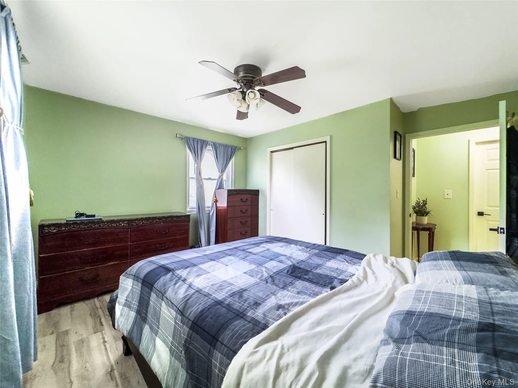 Wood-finish flooring, ceiling fan with integrated lighting, window with drapes, and a closet with bifold doors Wood-finish flooring, ceiling fan with integrated lighting, window with drapes, and a closet with bifold doors