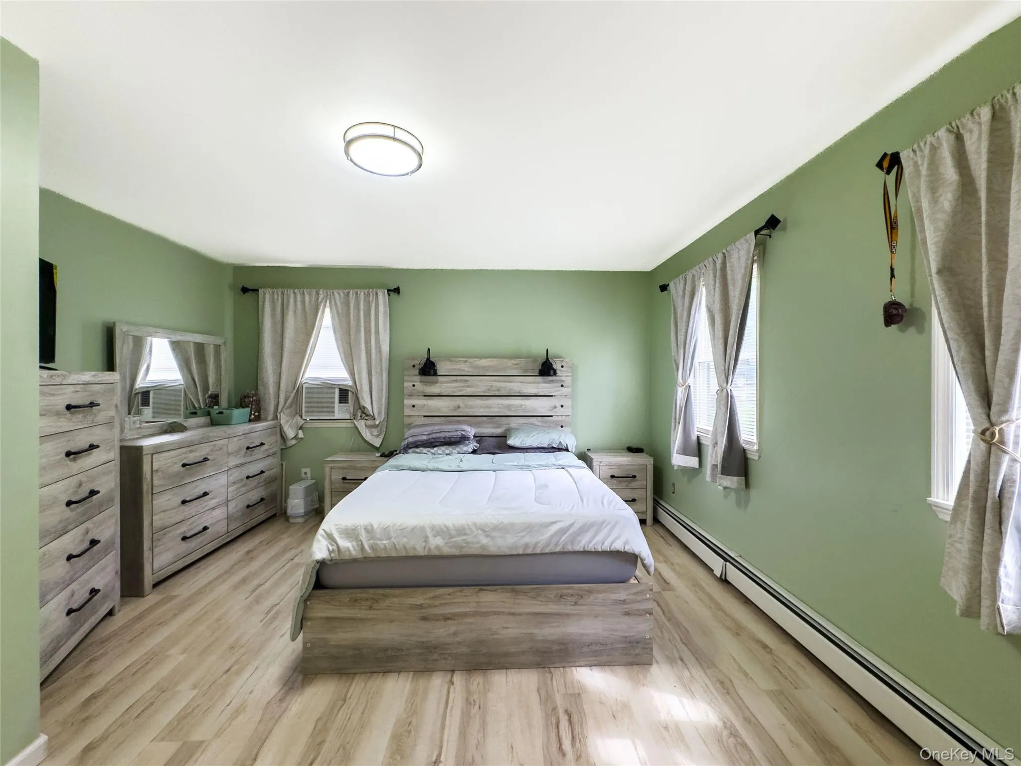 Spacious primary bedroom featuring soft sage-green walls, wood-finish flooring throughout, and abundant natural light from multiple windows with linen curtains Spacious primary bedroom featuring soft sage-green walls, wood-finish flooring throughout, and abundant natural light from multiple windows with linen curtains