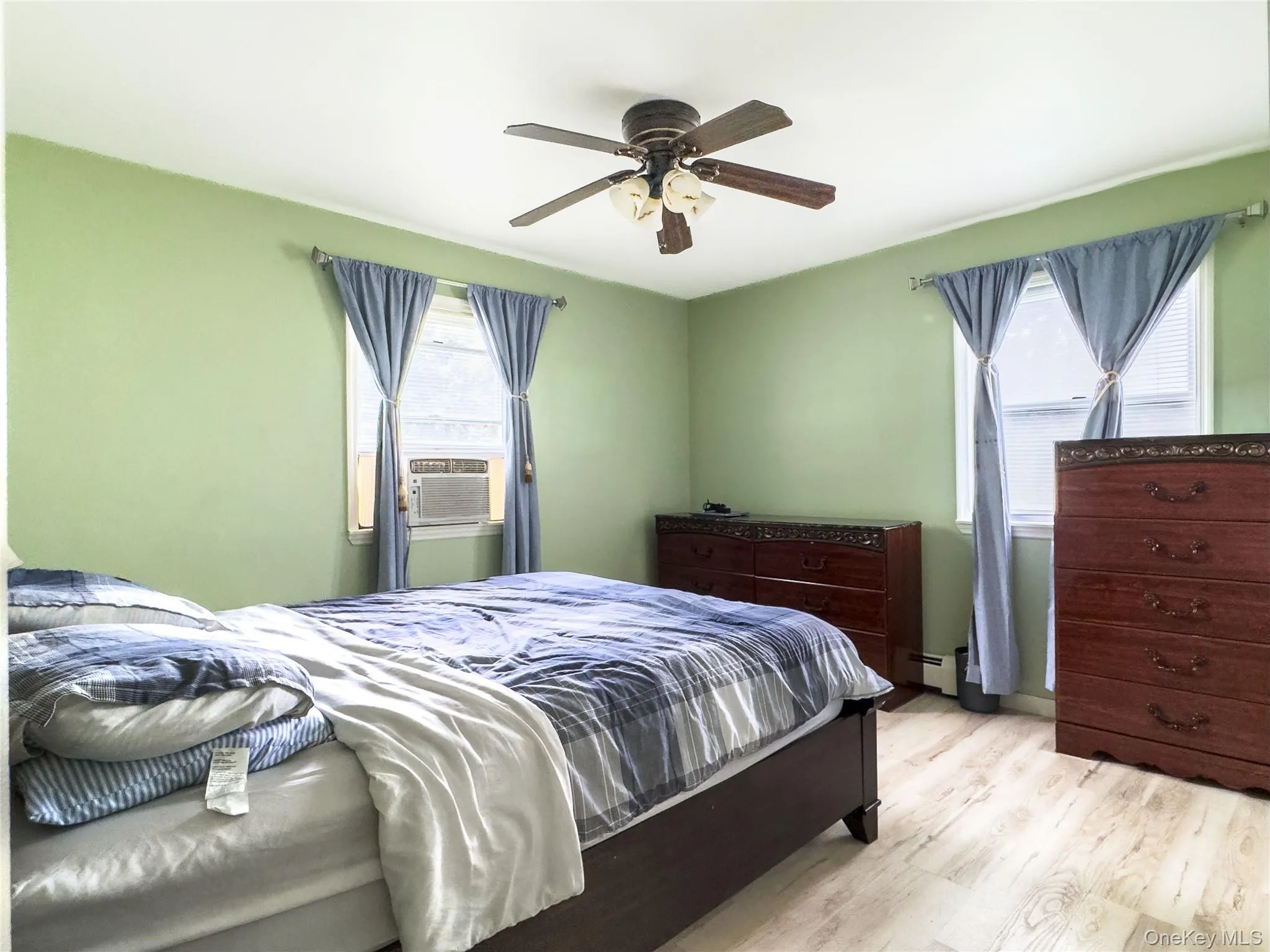 Bright bedroom with soft green walls, wood-finish flooring, and dual windows with blue drapery Bright bedroom with soft green walls, wood-finish flooring, and dual windows with blue drapery