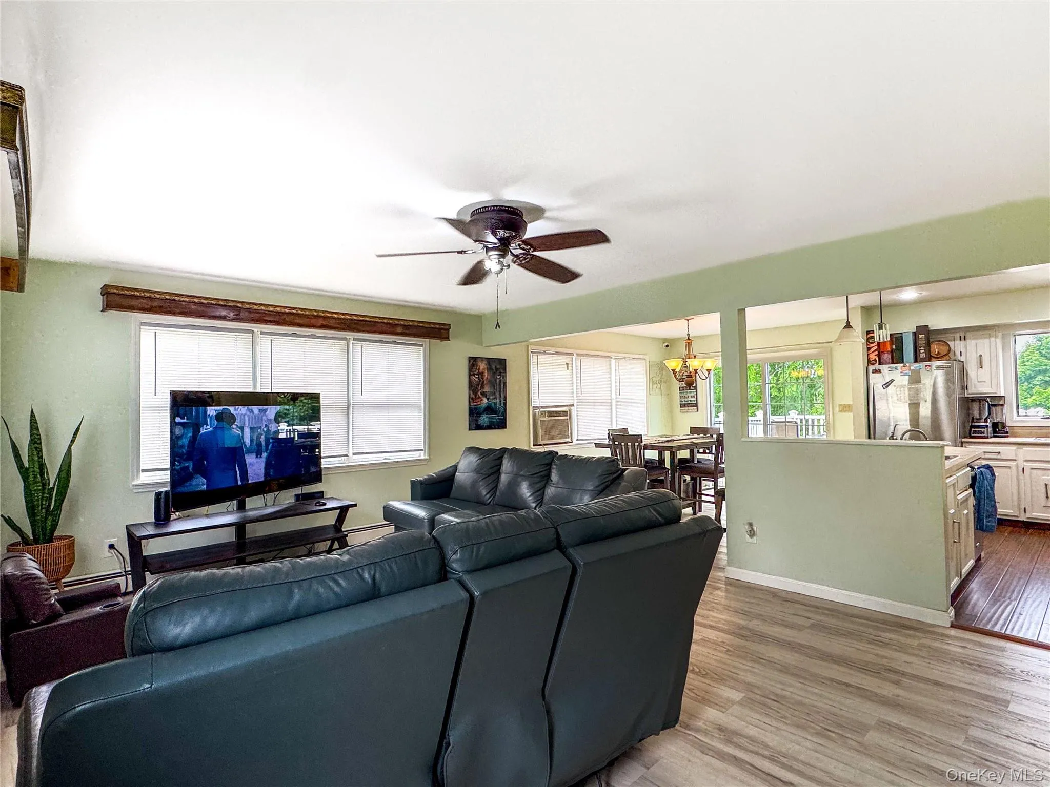 Open-concept living area with wood-finish flooring, ceiling fan with pull-chain controls, and large windows providing natural light Open-concept living area with wood-finish flooring, ceiling fan with pull-chain controls, and large windows providing natural light