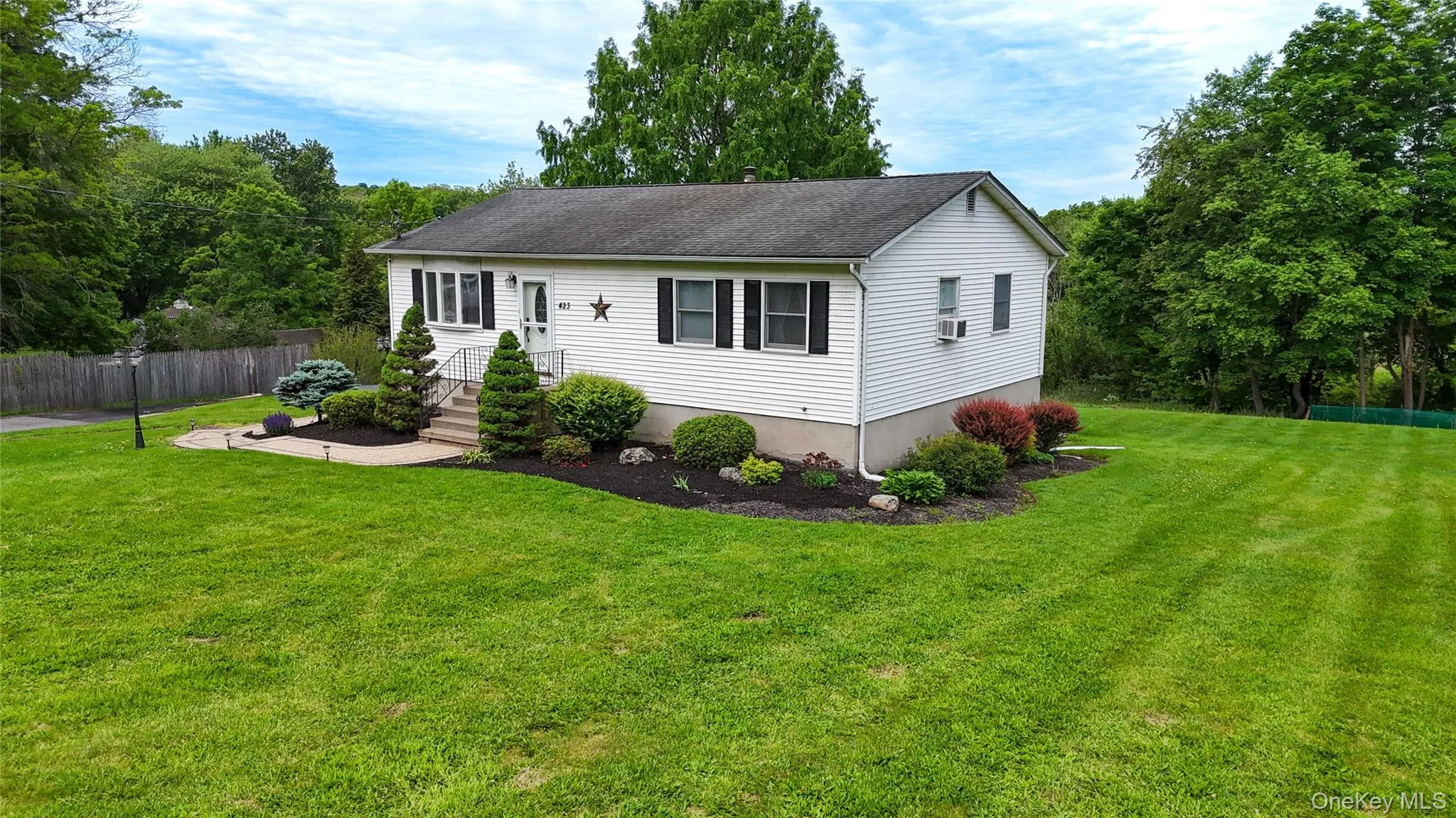White siding exterior with dark shutters, front entry with stone steps and handrails, landscaped beds with diverse plantings, large grassy lawn area, and mature perimeter trees White siding exterior with dark shutters, front entry with stone steps and handrails, landscaped beds with diverse plantings, large grassy lawn area, and mature perimeter trees