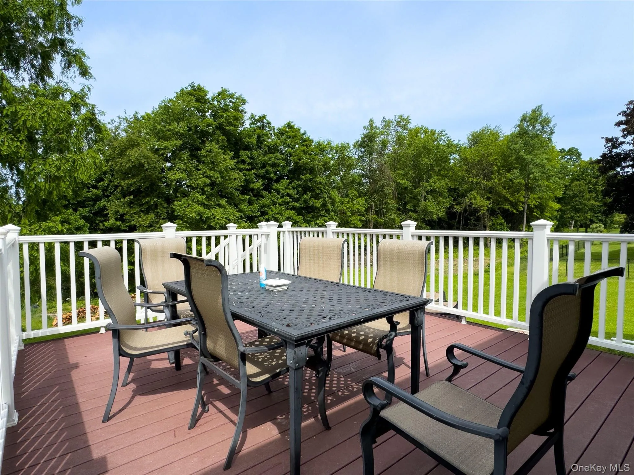 Expansive composite deck featuring composite decking with a black metal dining table and seatings, white vinyl railings with decorative balusters, and panoramic vistas of mature landscaping and tree line Expansive composite deck featuring composite decking with a black metal dining table and seatings, white vinyl railings with decorative balusters, and panoramic vistas of mature landscaping and tree line