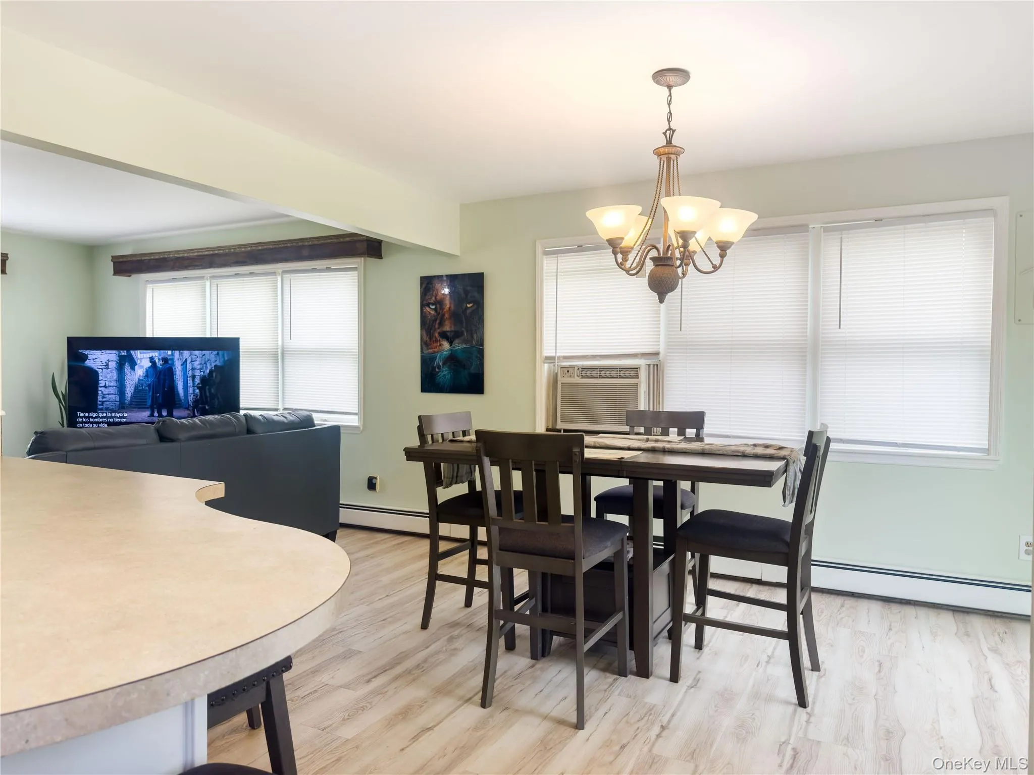Open-concept living and dining area featuring wood-finish flooring throughout, a curved counter island with seating, a dark wood dining table with matching chairs, large windows with cellular shades providing natural light, a brass chandelier with multiple arms, and a wall-mounted air conditioning unit Open-concept living and dining area featuring wood-finish flooring throughout, a curved counter island with seating, a dark wood dining table with matching chairs, large windows with cellular shades providing natural light, a brass chandelier with multiple arms, and a wall-mounted air conditioning unit