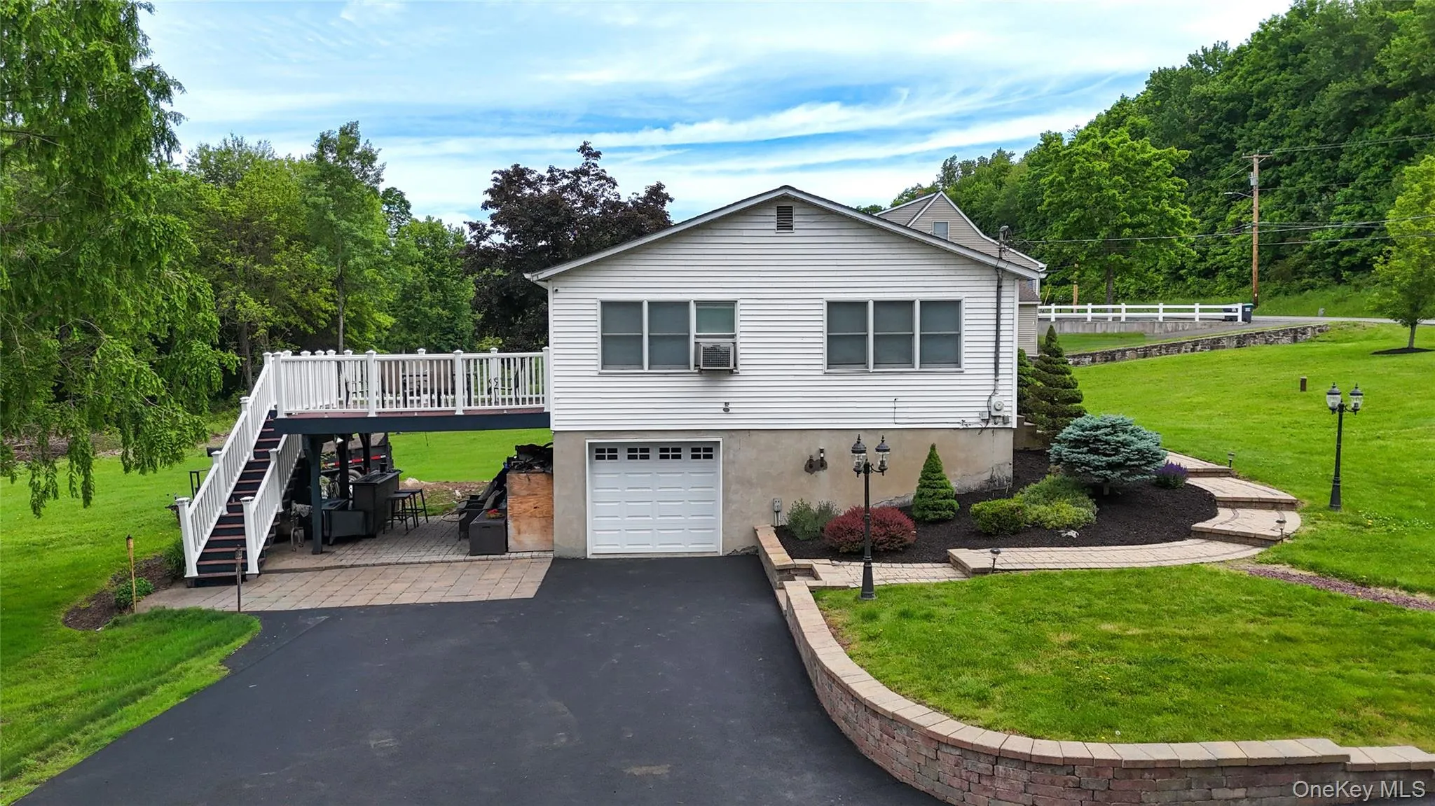 Elevated home featuring a clean white exterior with dual garage doors, expansive asphalt driveway, and manicured grounds with mature landscaping Elevated home featuring a clean white exterior with dual garage doors, expansive asphalt driveway, and manicured grounds with mature landscaping