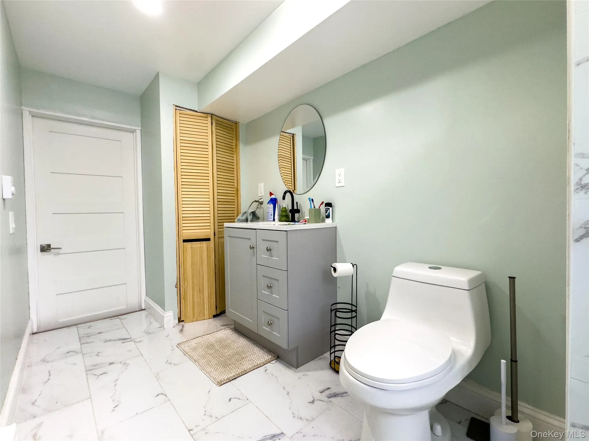 Modern primary bathroom featuring soft sage green walls, polished marble-look tile flooring, a gray vanity with integrated sink and ample storage, a contemporary toilet with elongated bowl, and circular mirror above the vanity Modern primary bathroom featuring soft sage green walls, polished marble-look tile flooring, a gray vanity with integrated sink and ample storage, a contemporary toilet with elongated bowl, and circular mirror above the vanity
