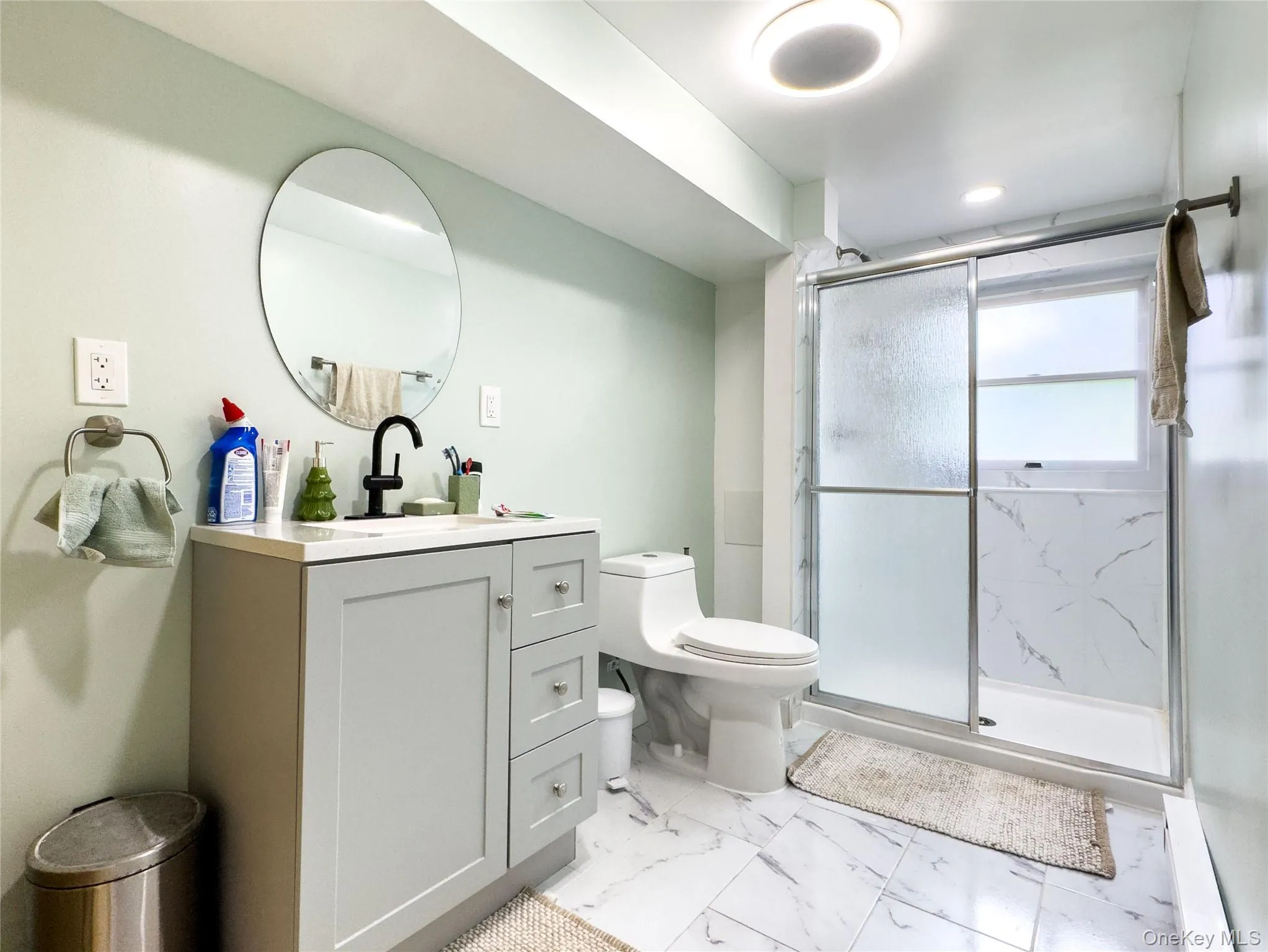 Modern primary bathroom featuring a white vanity with ample storage, polished stone-finish flooring, frameless glass shower enclosure with sliding doors, and a circular mirror with integrated lighting above the sink Modern primary bathroom featuring a white vanity with ample storage, polished stone-finish flooring, frameless glass shower enclosure with sliding doors, and a circular mirror with integrated lighting above the sink