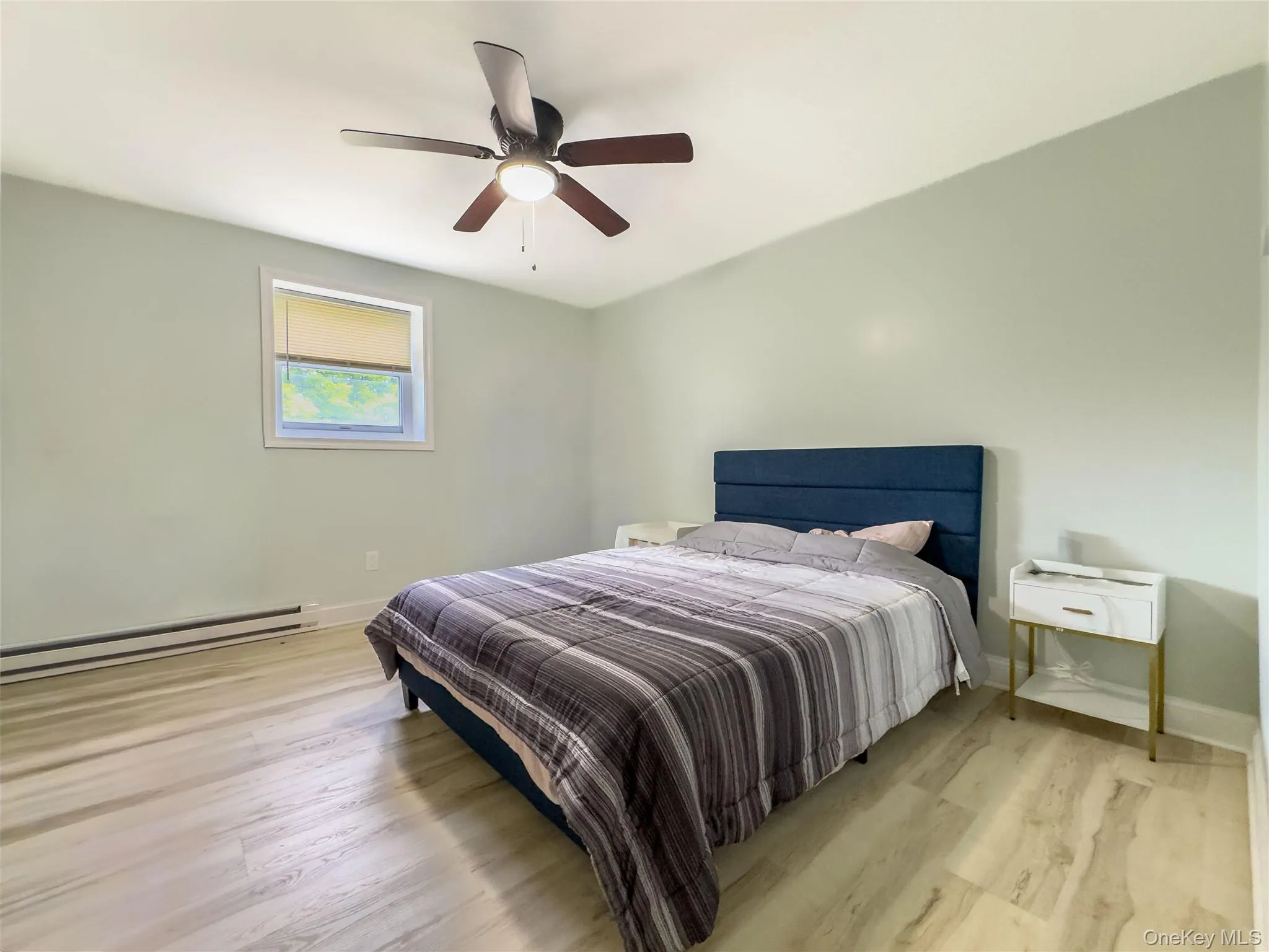 Bright bedroom featuring a wood-finish plank floor, soft green wall finish, large single-hung window with natural light, ceiling fan with integrated lighting, and a blue upholstered bed frame Bright bedroom featuring a wood-finish plank floor, soft green wall finish, large single-hung window with natural light, ceiling fan with integrated lighting, and a blue upholstered bed frame