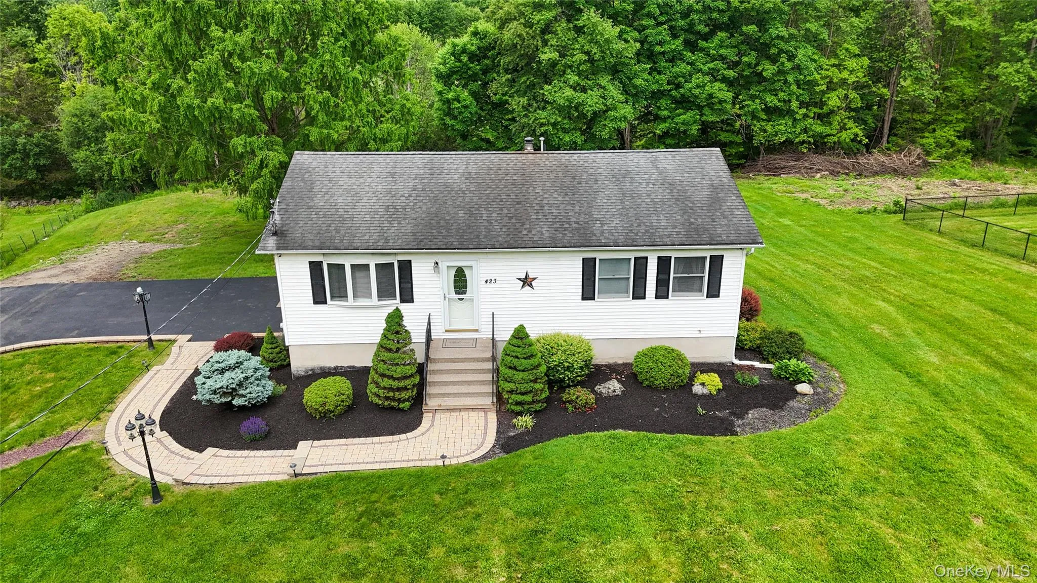 Charming ranch-style home featuring a freshly sealed asphalt driveway, manicured landscaping with symmetrical evergreen plantings, and a curved brick walkway leading to the entrance Charming ranch-style home featuring a freshly sealed asphalt driveway, manicured landscaping with symmetrical evergreen plantings, and a curved brick walkway leading to the entrance