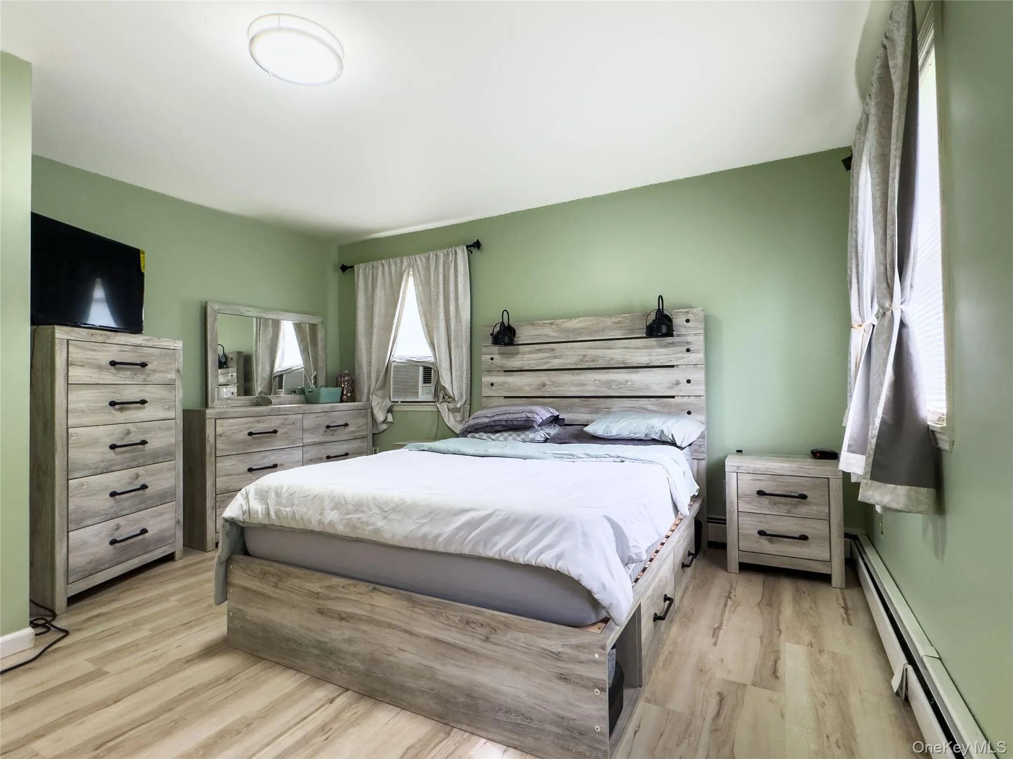 Primary Bedroom Suite with Soft Sage Green Walls, Wood-Finish Flooring, and Coordinated Wood Bedroom Furnishings Including Dresser and Nightstand Primary Bedroom Suite with Soft Sage Green Walls, Wood-Finish Flooring, and Coordinated Wood Bedroom Furnishings Including Dresser and Nightstand