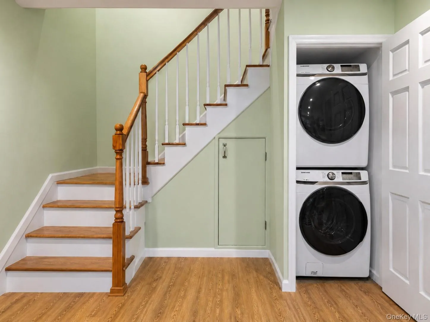 Wood-finish flooring, staircase with wood handrail, white balusters, and wood treads, built-in stacked laundry appliances, and a solid panel door Wood-finish flooring, staircase with wood handrail, white balusters, and wood treads, built-in stacked laundry appliances, and a solid panel door